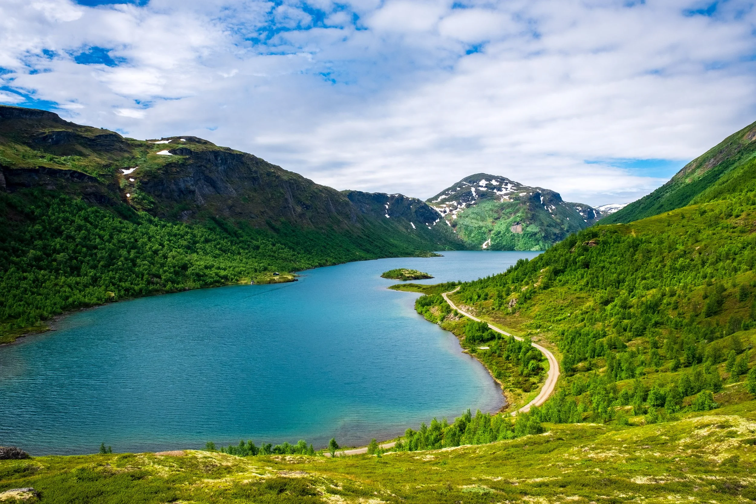 En innsjø omgitt av grønne fjell med snø på toppene, en liten vei langs vannet og en blå himmel med skyer.