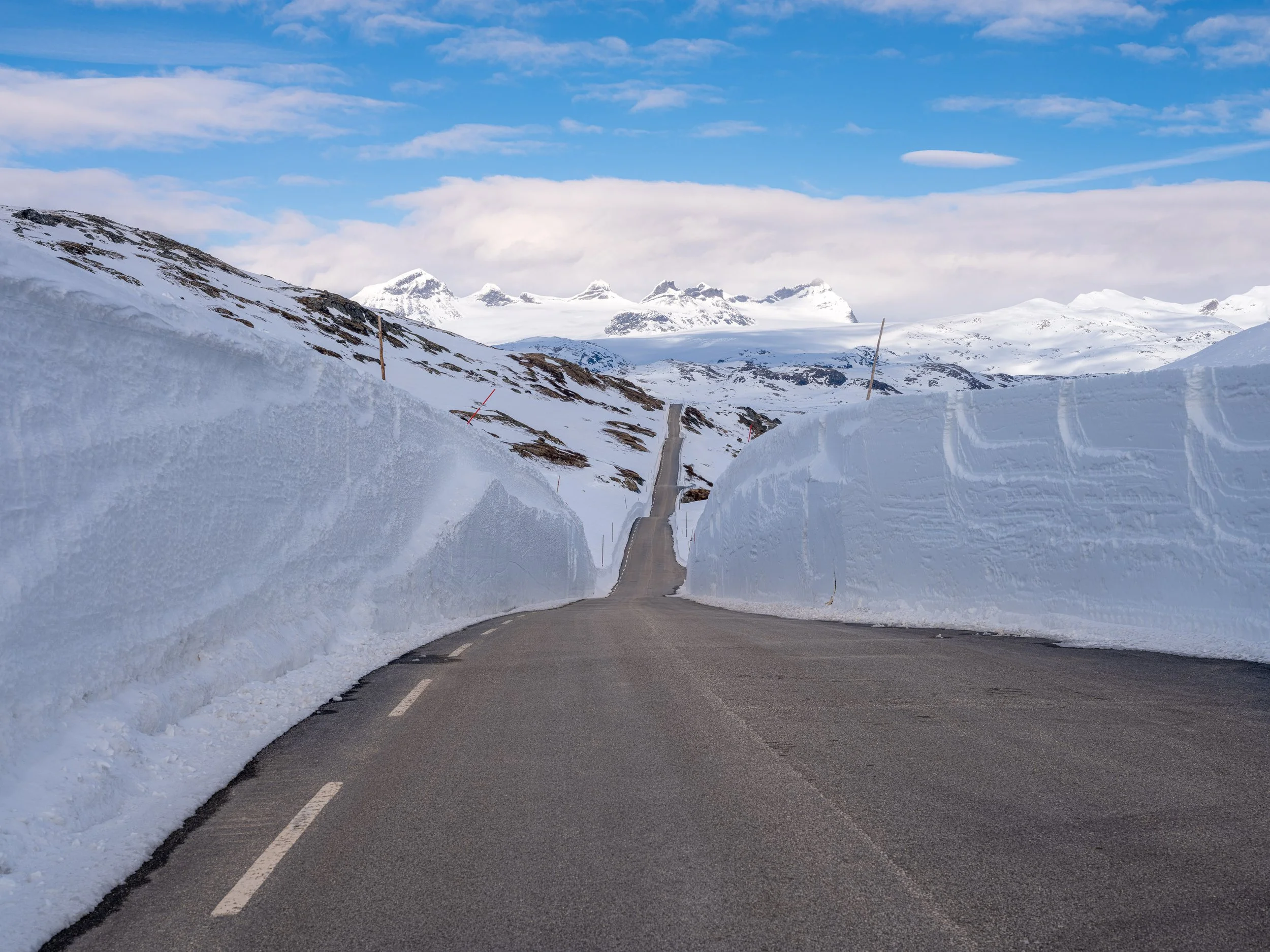 Sognefjellsvegen med høye snøvegger på begge sider, omgitt av snødekte fjell og en blå himmel.