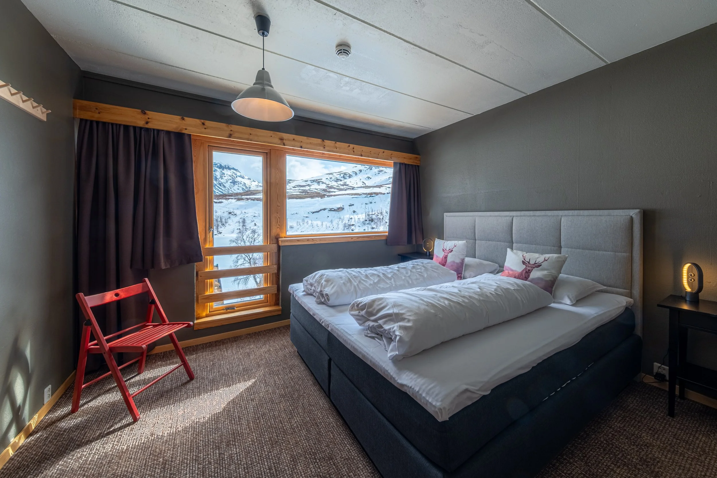 Innendørs bilde av dobbeltrom med dobbeltseng, store vindu som viser fjell og snø lett snø, mørke gardiner, et liten svart nattbord med en lysende lampe, og en rød stol