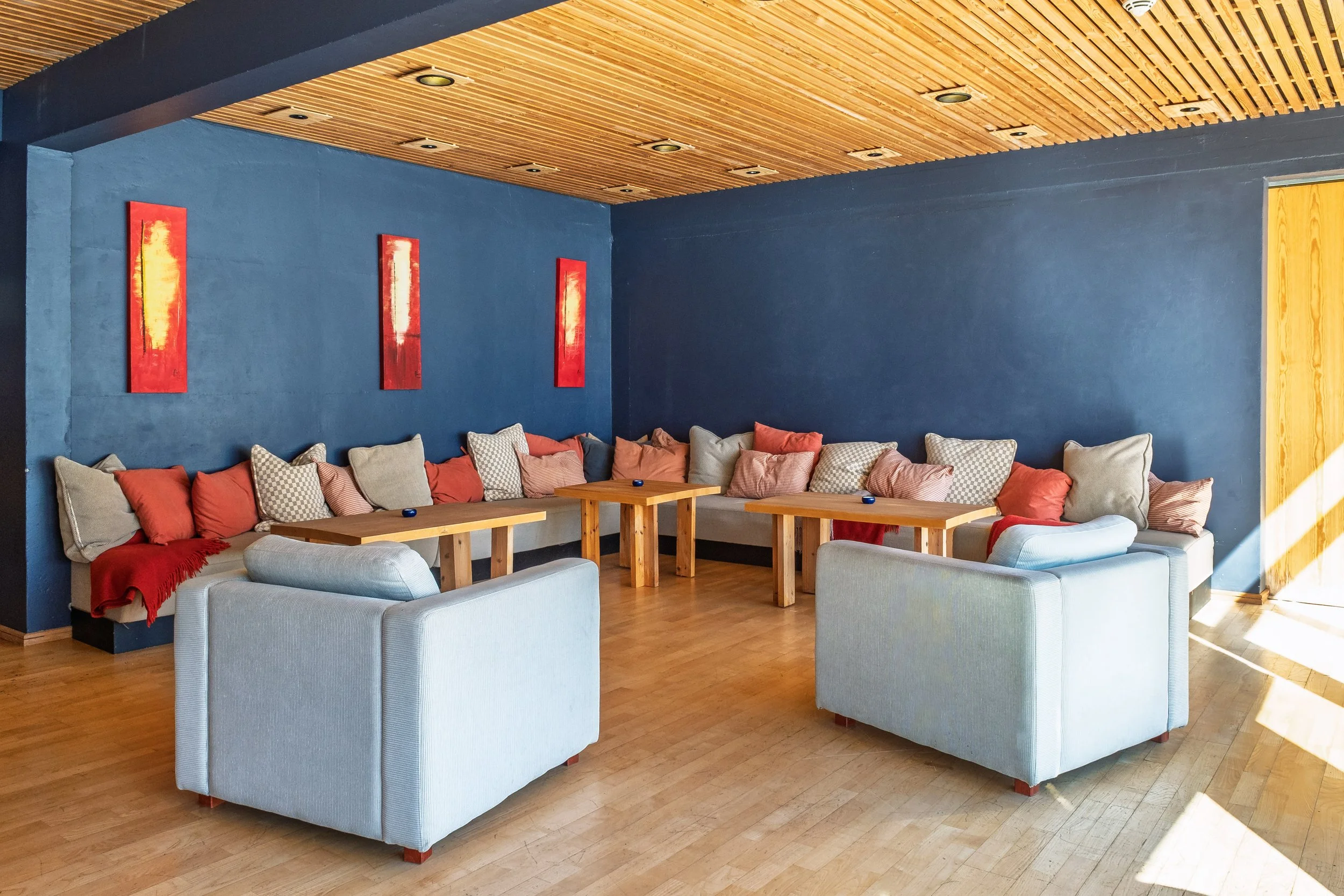 Stue med blå vegg, beige sofaer og trebord. Tre malerier med varme nyanser på veggen, lys trepanel i taket, og solskinn som skinner inn gjennom vindu på høyre side.