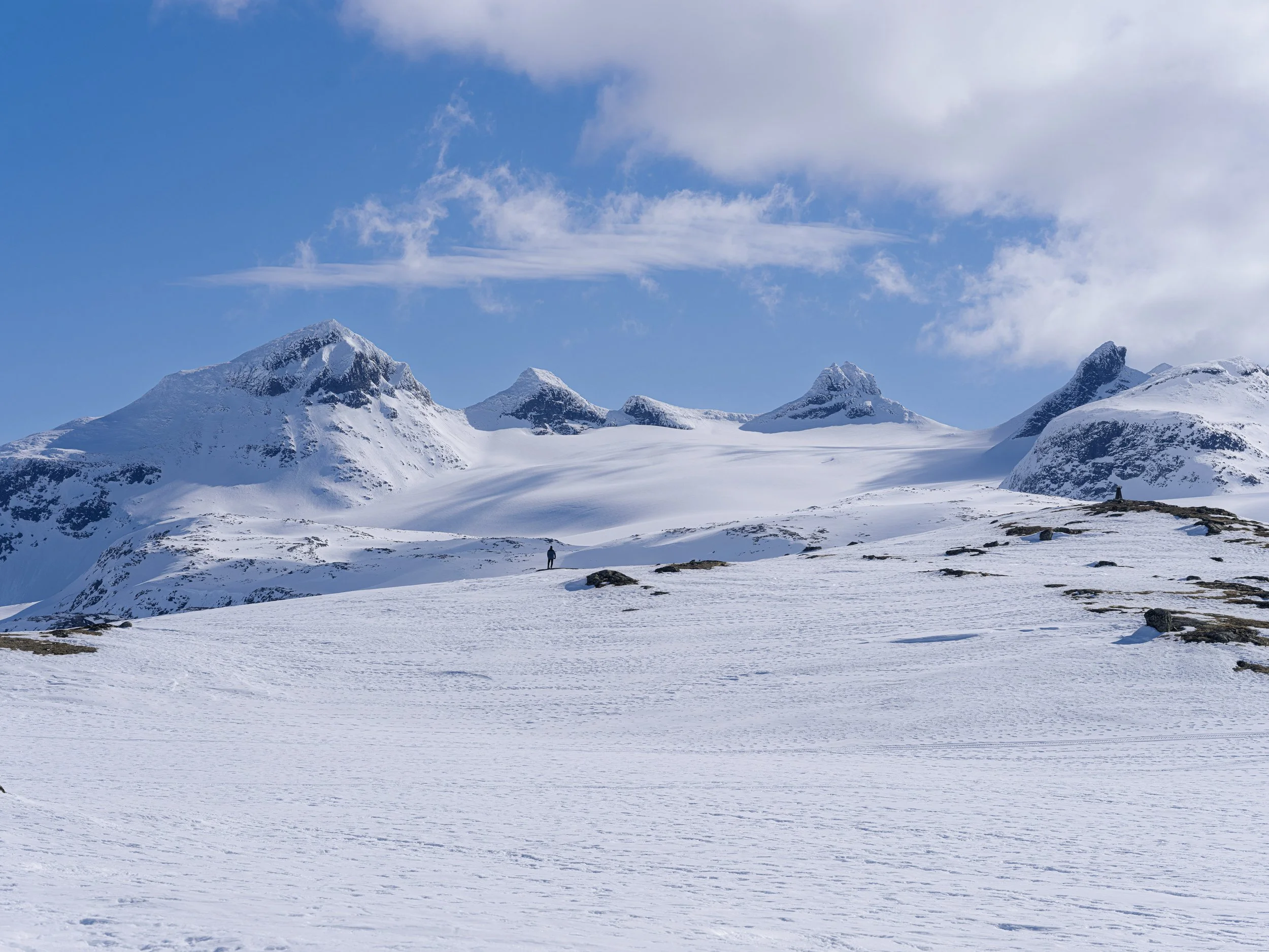 Snødekt fjelllandskap med flere topper og en ensom person som går i snøen under en blå himmel med spredte skyer.