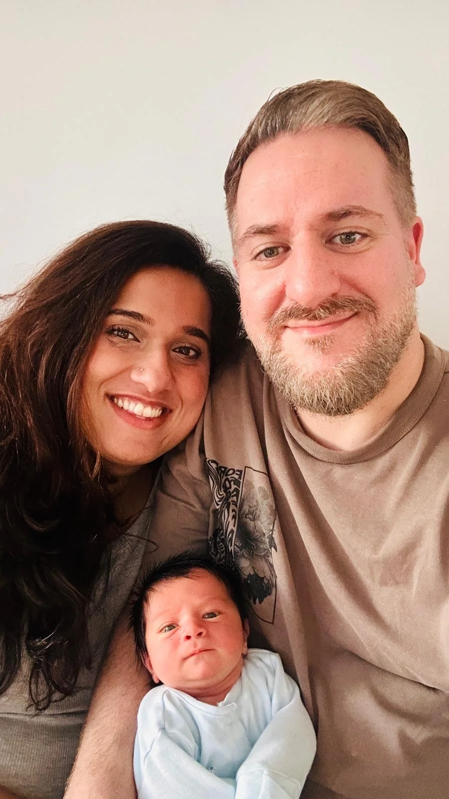 A smiling couple taking a selfie with their newborn baby against a plain white wall. After having a positive birth experience using hypnobirthing tools.