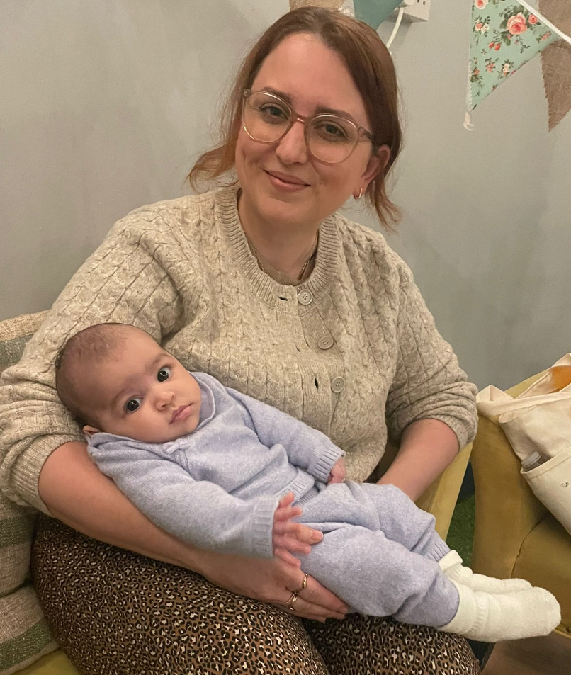 A woman with glasses and a beige sweater smiling while holding a baby in a light blue outfit, sitting on her lap in a cozy indoor setting with floral decorations.