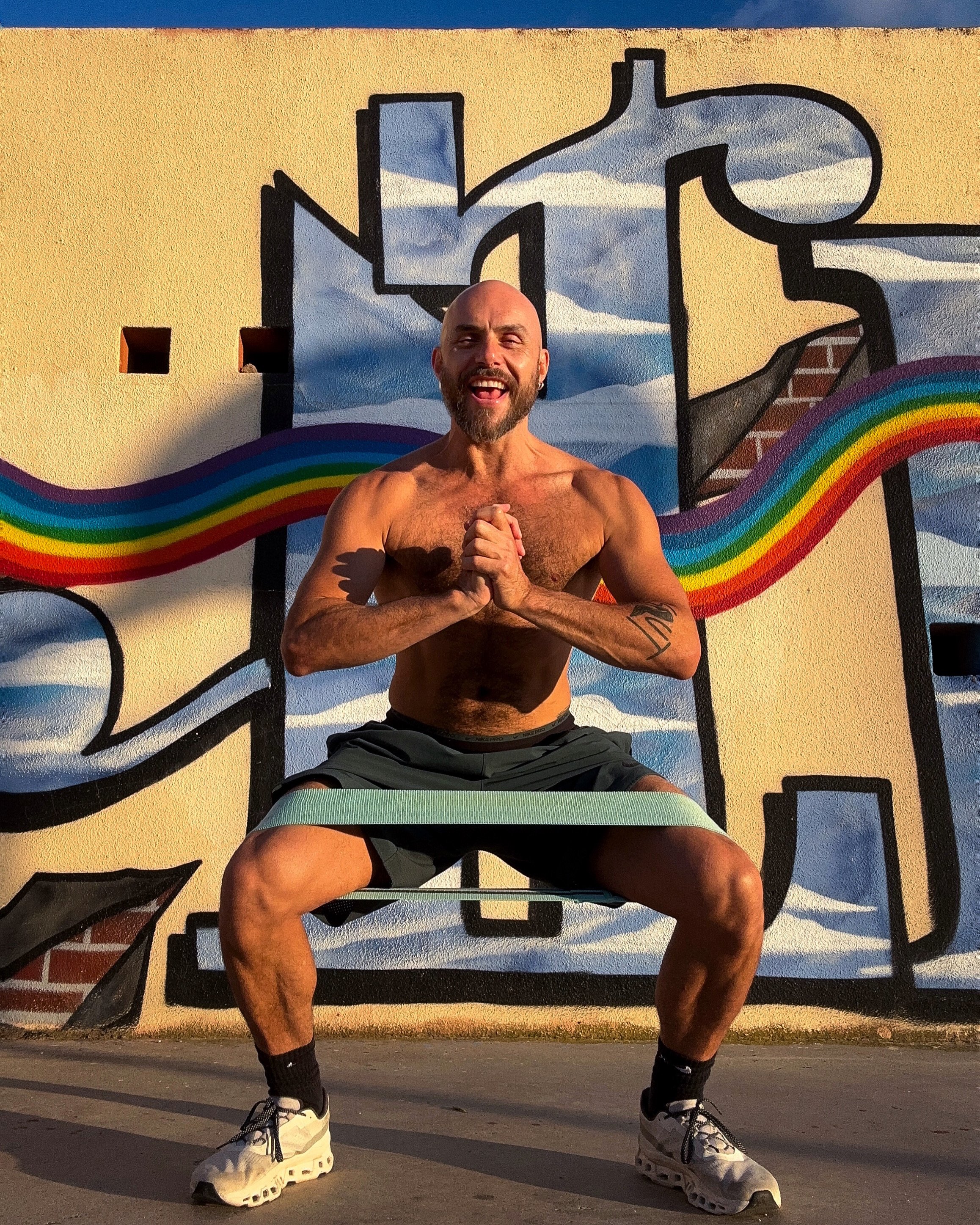 Man exercising outdoors with resistance band in front of colorful graffiti wall, wearing shorts and shoes.