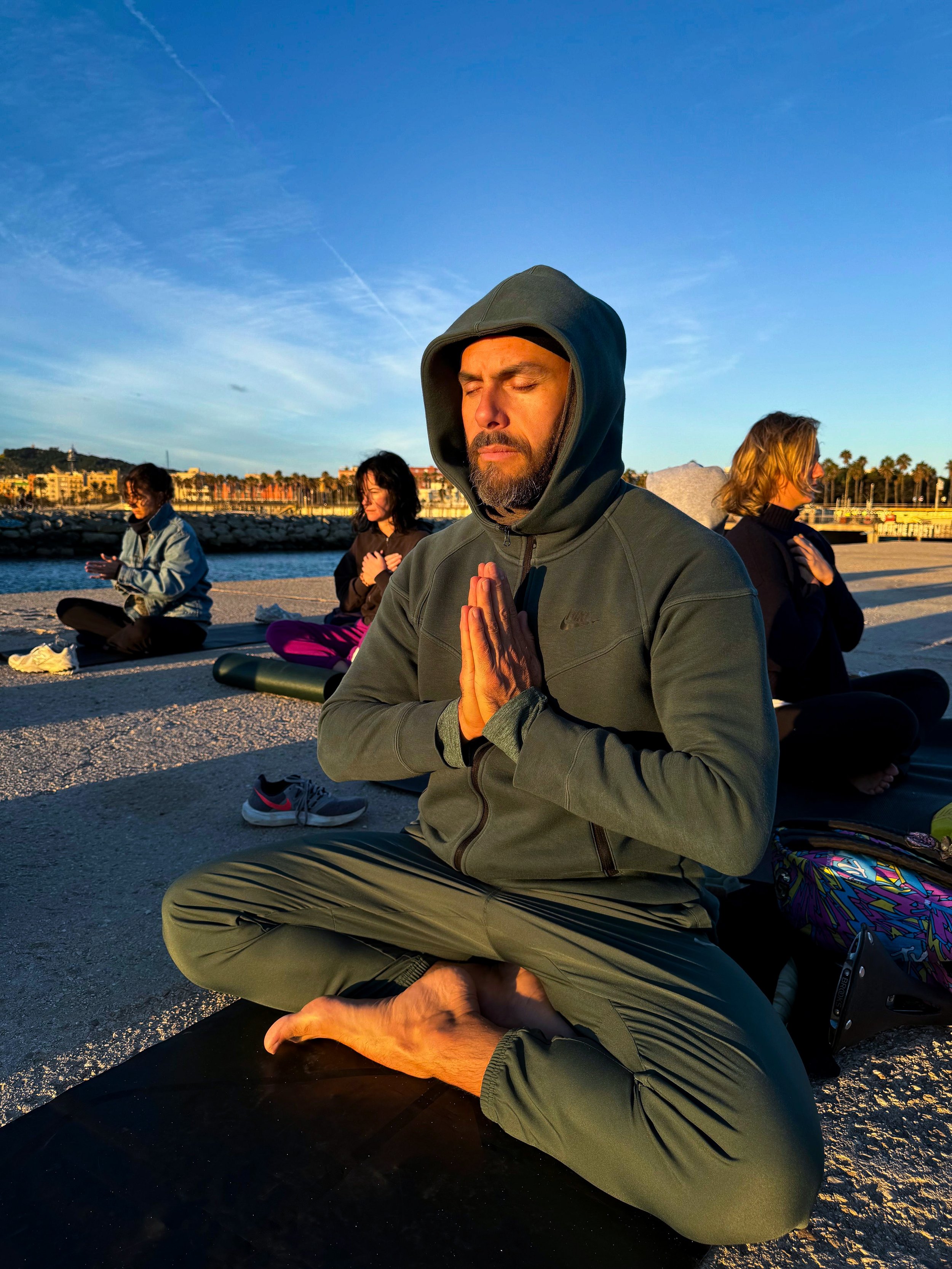 A person meditating on a beach at sunset with several others in the background, all sitting cross-legged with hands in prayer position. The main person is wearing a green hoodie.