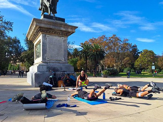 Group exercising outdoors on yoga mats near a large statue in a park, with trees and clear sky in the background.