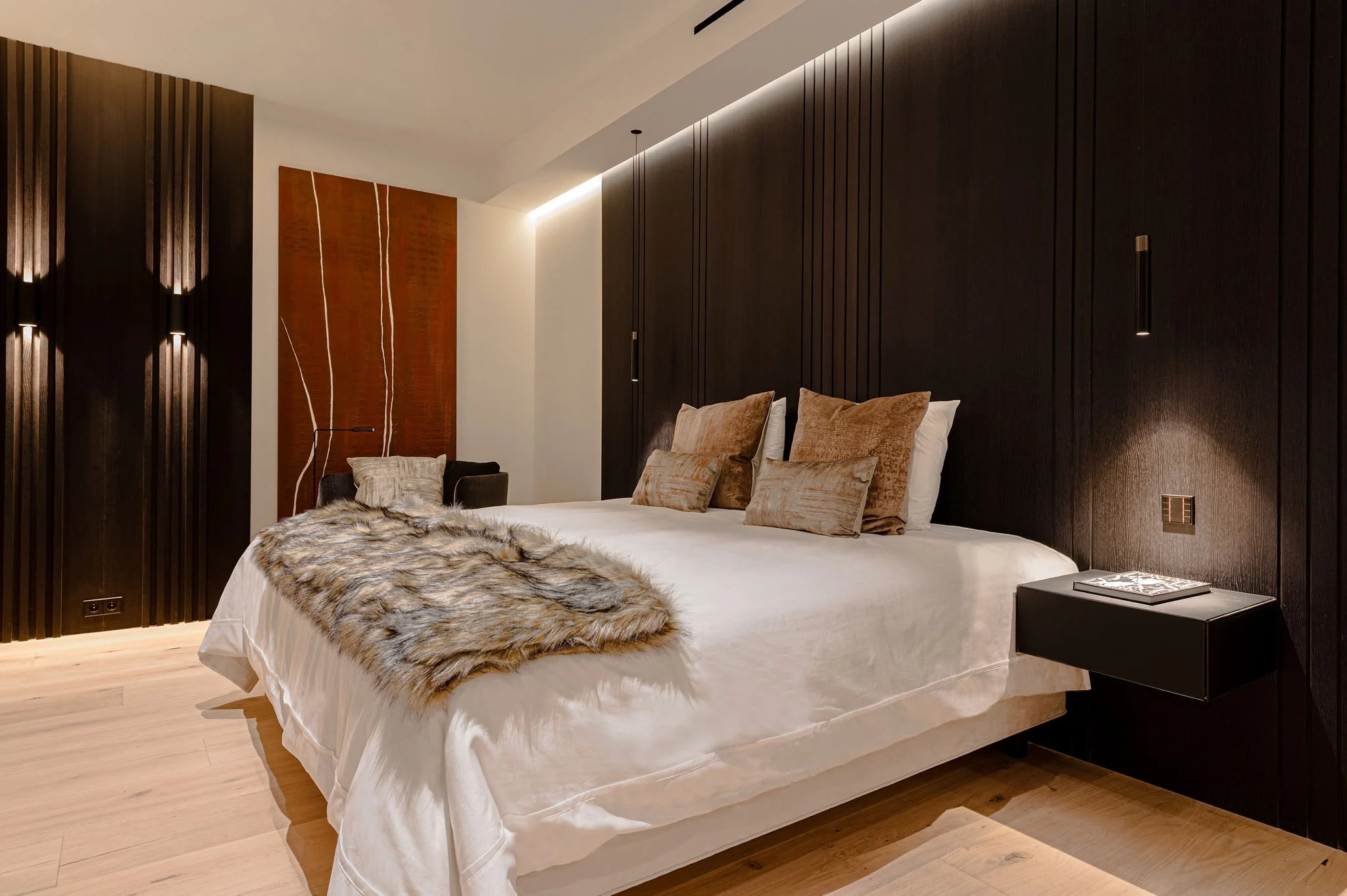 Dormitorio con paredes de madera oscura, una cama con ropa blanca y varios cojines de tonos beige y marrón, una manta de pelo en la cama, una lámpara negra sobre una mesita y arte en la pared