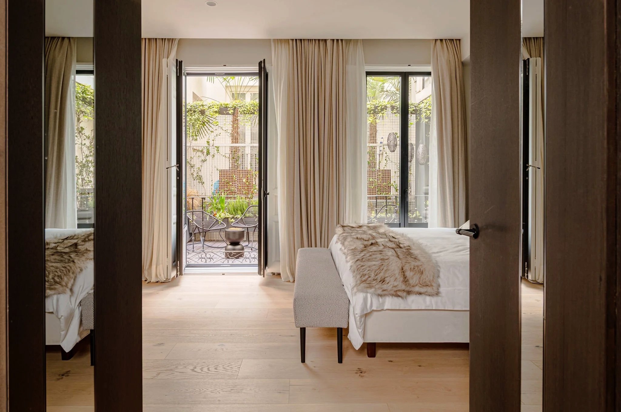 Dormitorio con puertas abiertas a un balcón y cortinas beige, con camas con mantas de piel artificial, vista a plantas y árboles en el balcón.