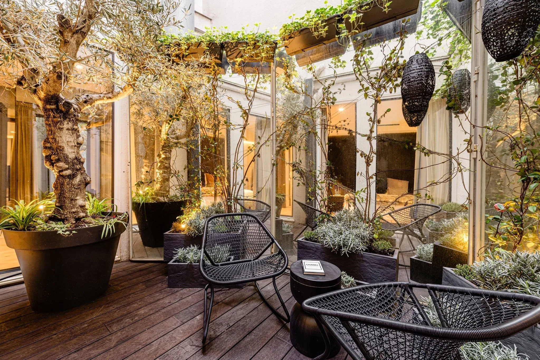 Un patio con plantas y árboles en macetas grandes, con muebles de mimbre negro y una libreta en una mesa redonda negra, rodeado de paredes de vidrio con cortinas y luz cálida.