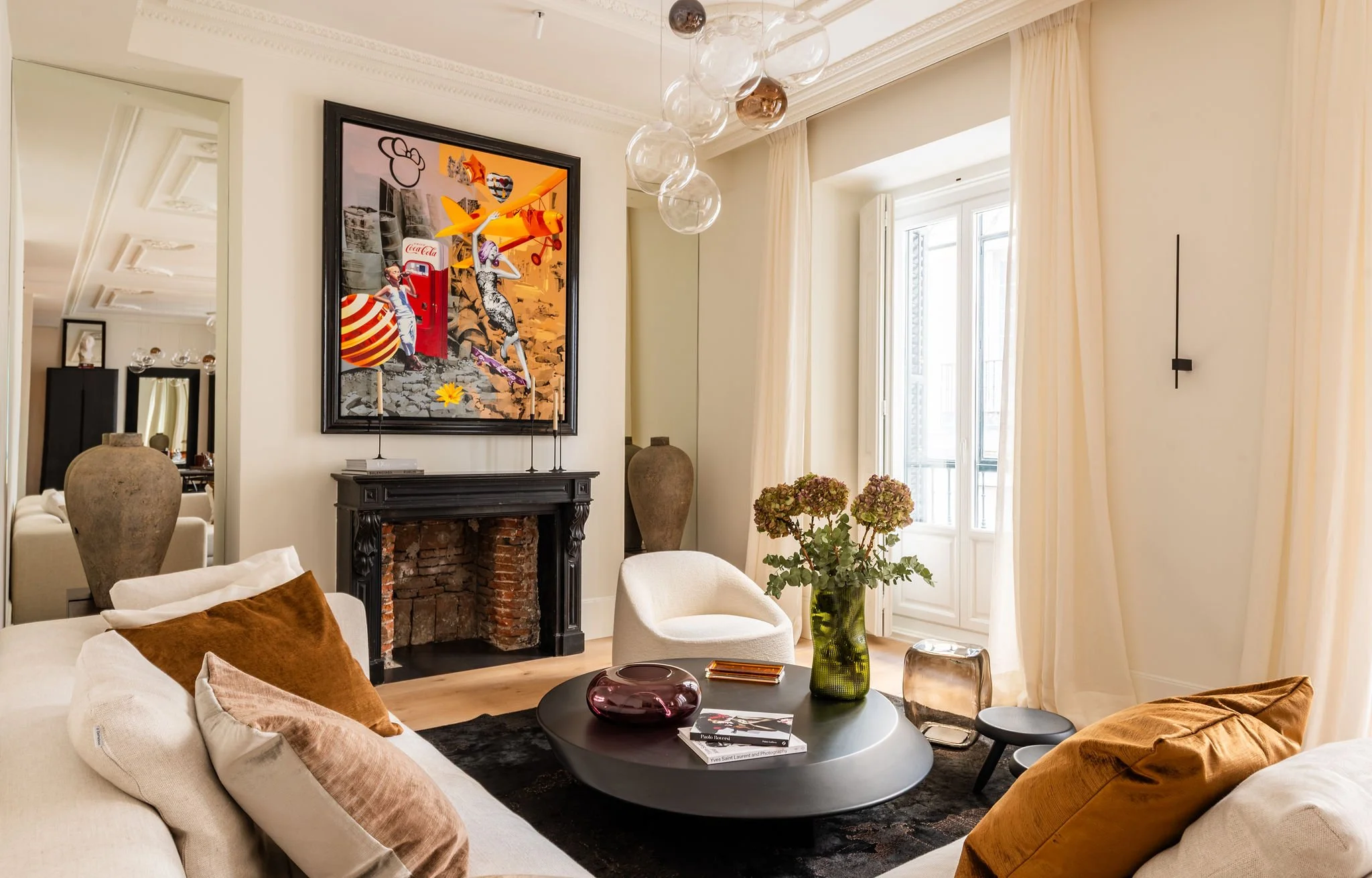 Sala de estar con sillones blancos y cobijas marrón, mesa de centro negra con libros y decoraciones, planta en jarrón verde y ventanas grandes con cortinas claras, chimenea y una pintura moderna en la pared.