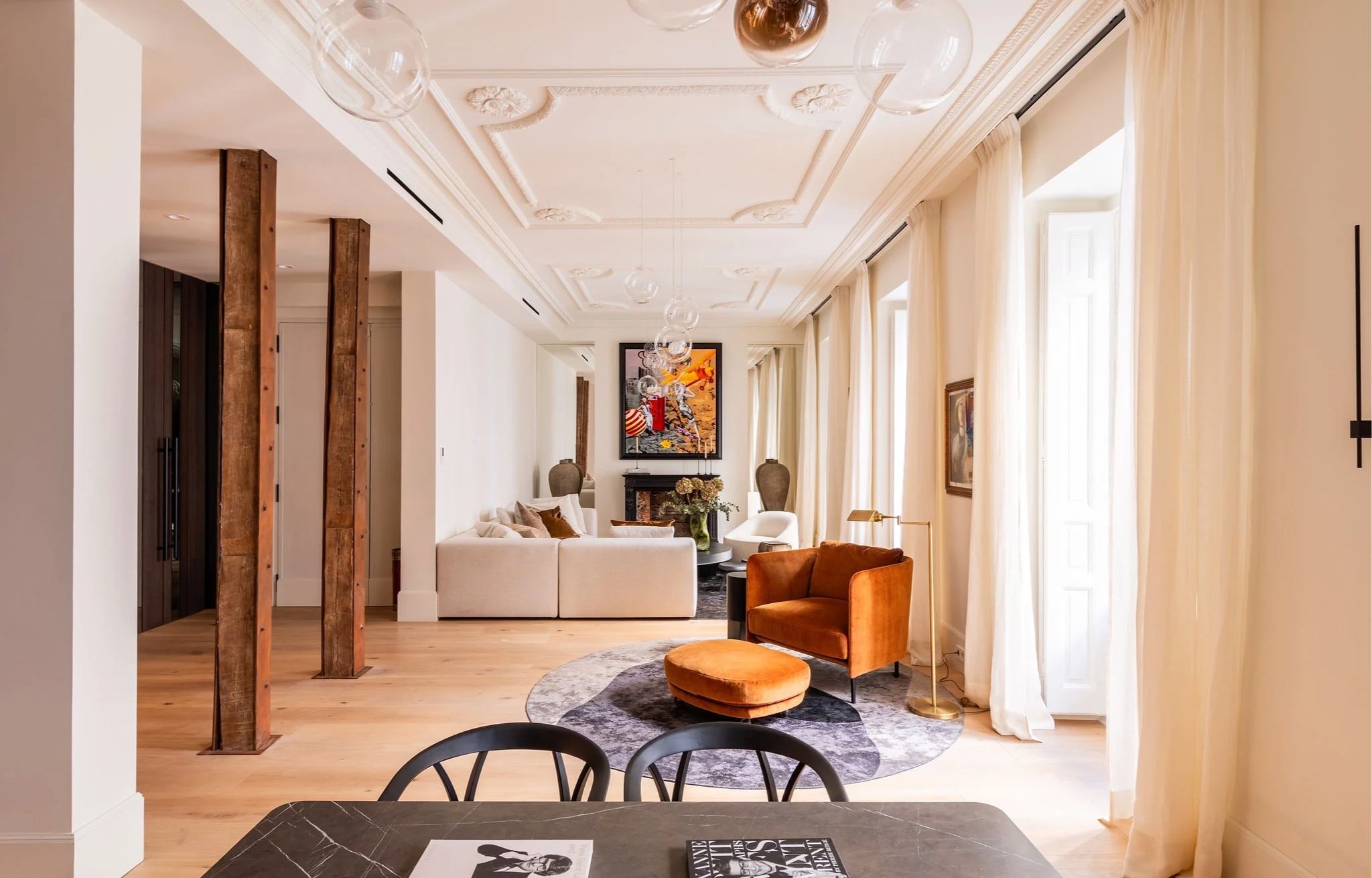 Sala de estar con sofá beige, sillón naranja y mesa de centro con flores, lámpara dorada y cuadros en las paredes, ventanas con cortinas blancas y techo decorado con molduras y lámparas colgantes de vidrio.