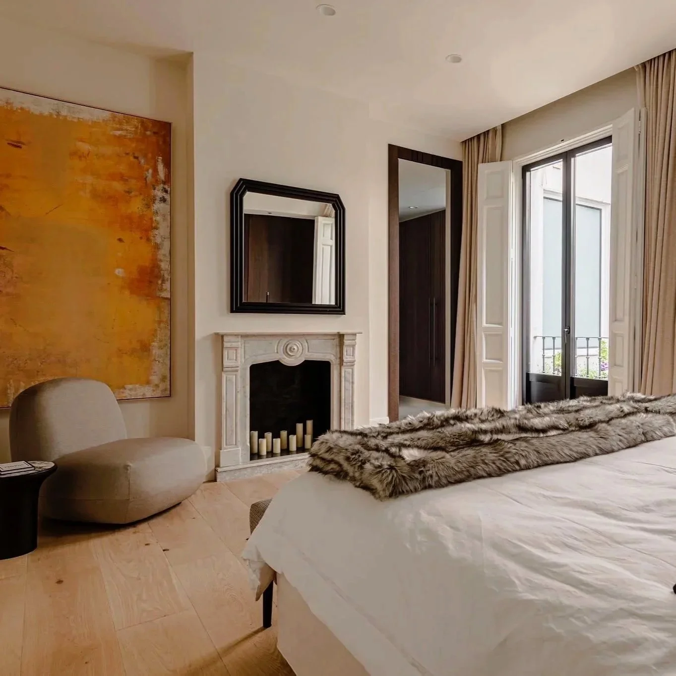 Una habitación con cama, chimenea, espejo, silla y ventana con cortinas beige que da a un balcón con barandilla.