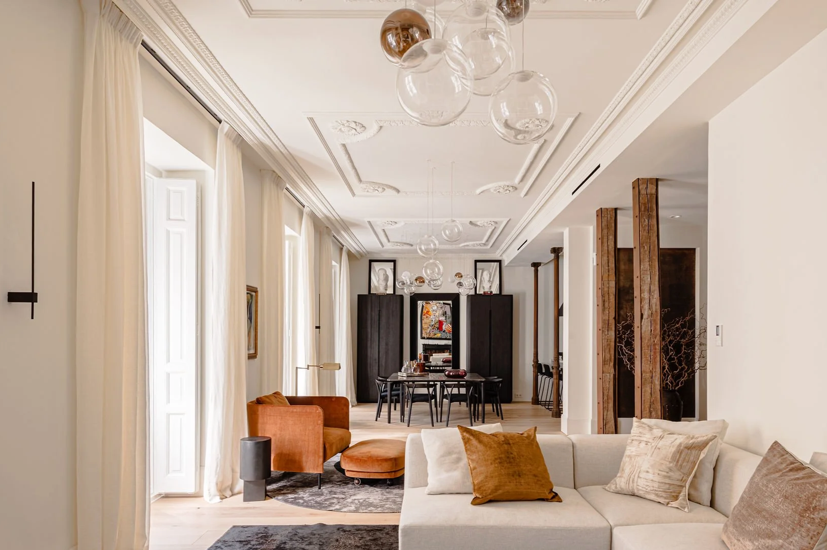 Sala elegante con techo decorativo, cortinas blancas, sillones en tonos tierra, cojines beige, una mesa de comedor, e iluminacion moderna con lámparas de vidrio.