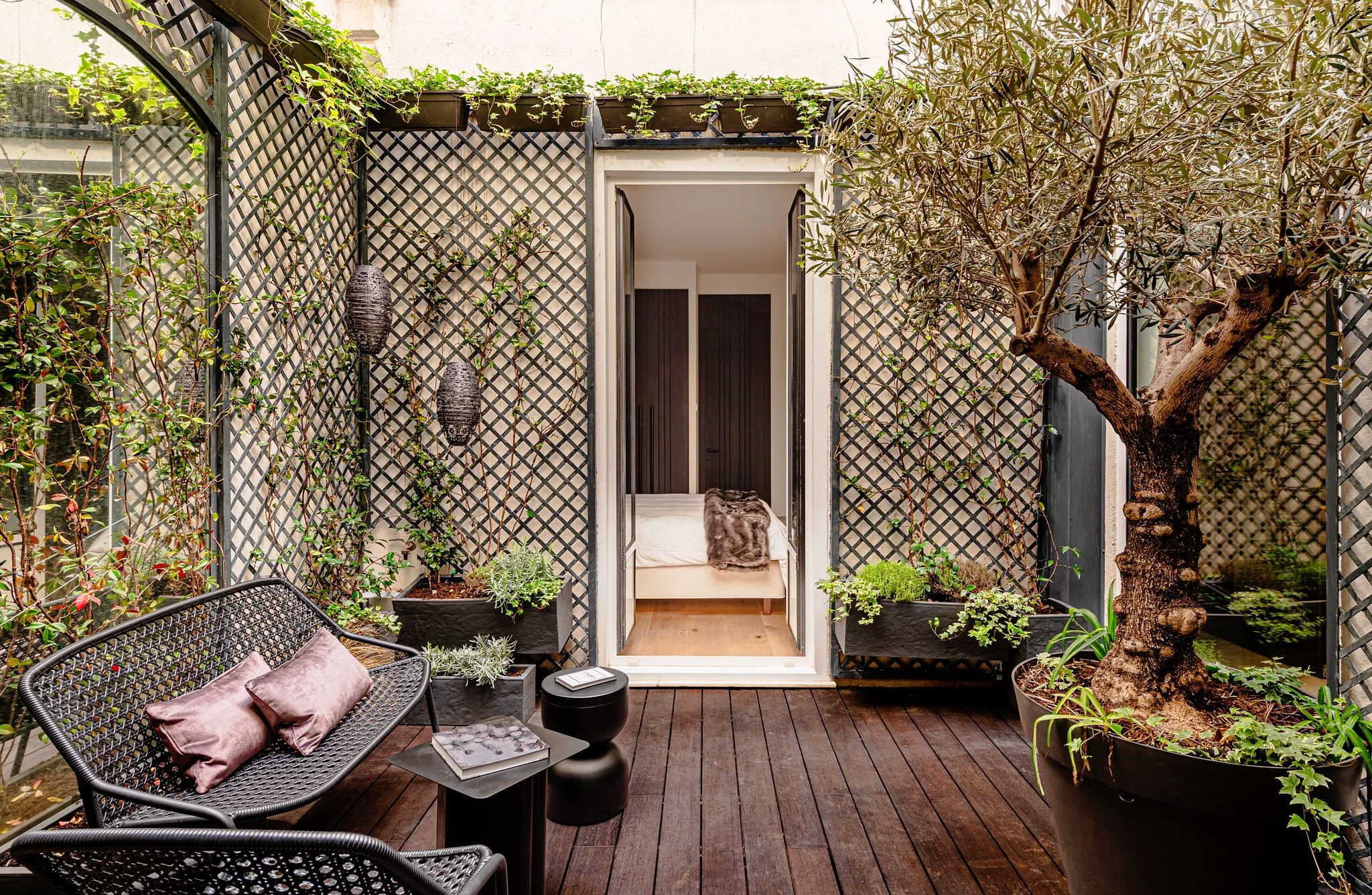 Rincón de patio con plantas, muebles de exterior y vista a un interior con cama y puertas negras.