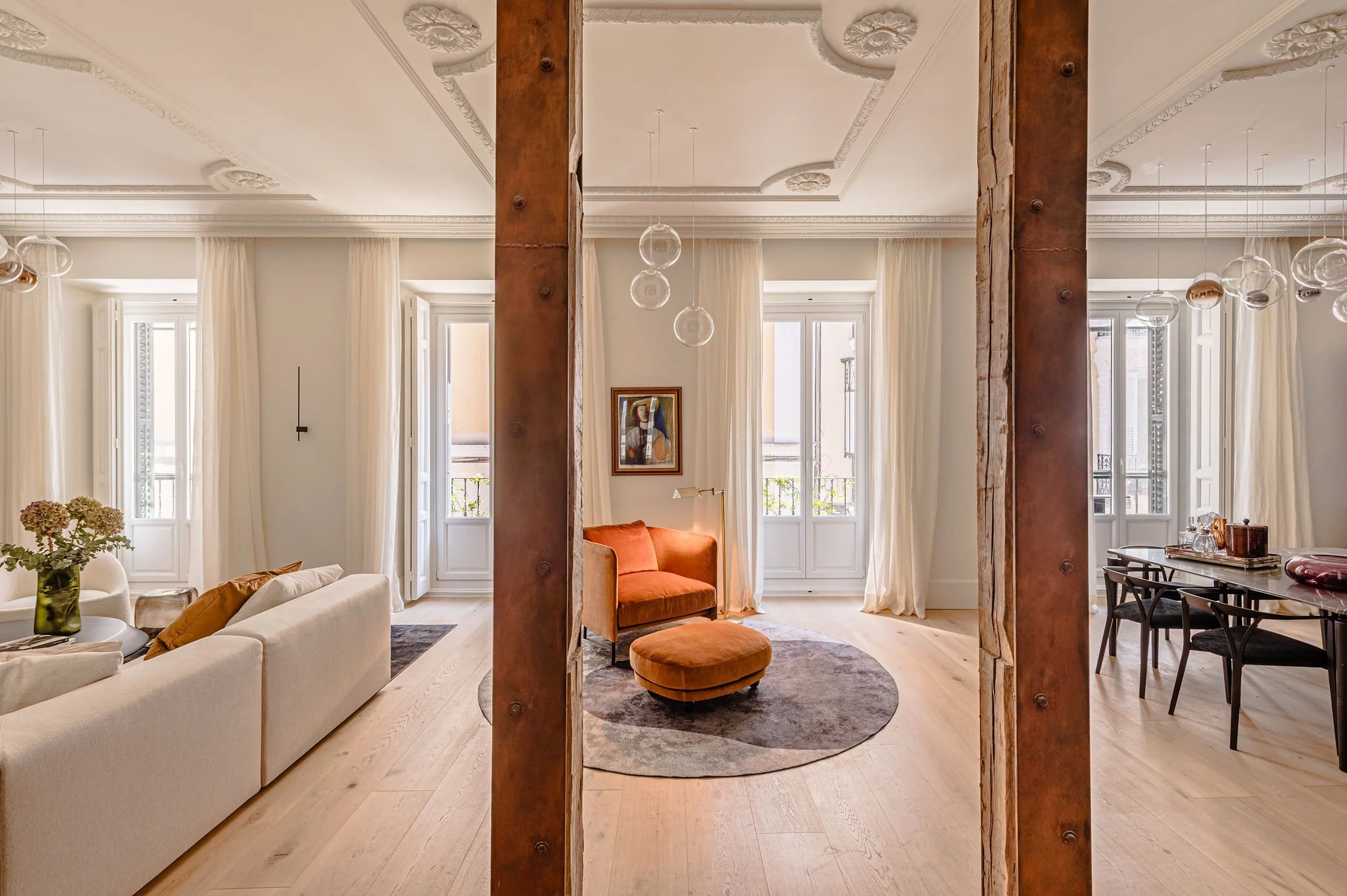 Vista interior de una sala de estar y comedor con grandes ventanas, cortinas blancas, sillón beige, sillón naranja y mesa de comedor con sillas negras, en un espacio decorado con techo con molduras y techos altos.