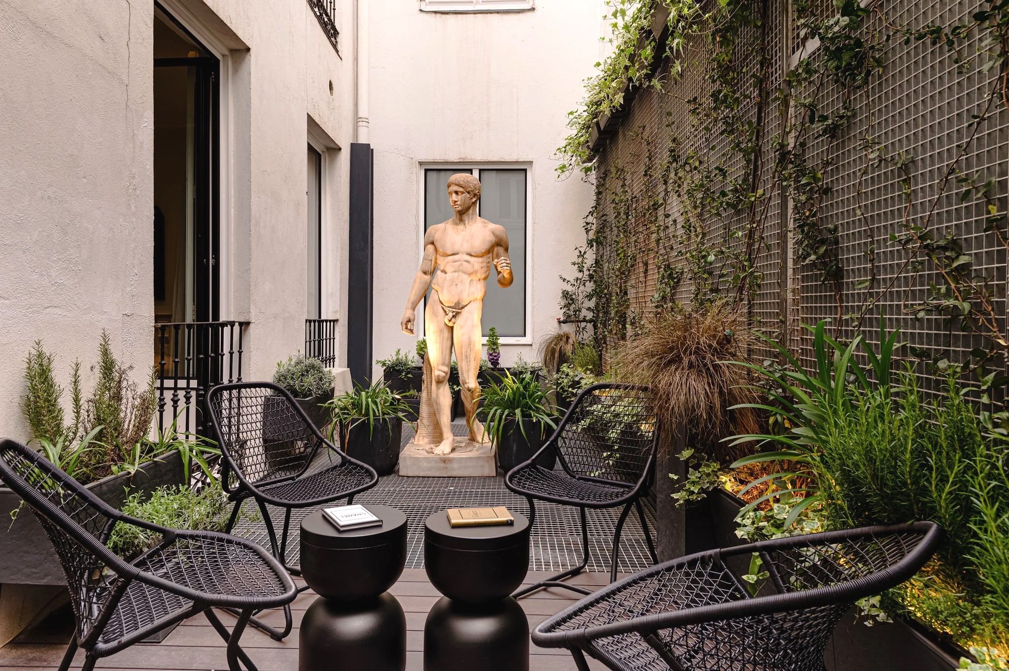 Una nueva escultura de un hombre desnudo en un patio rodeado de plantas y sillas de mimbre negras.