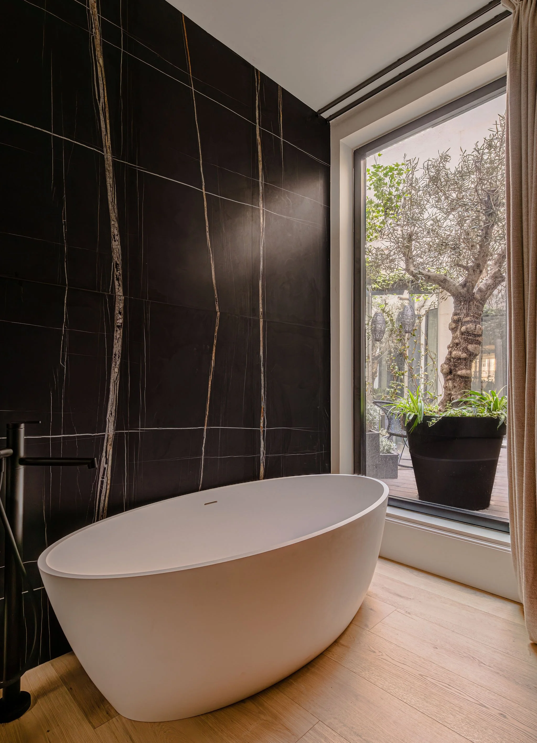Tina de baño de pie en un baño moderno con pared de azulejos negra y una ventana grande con vista a un árbol, plantas y un patio.