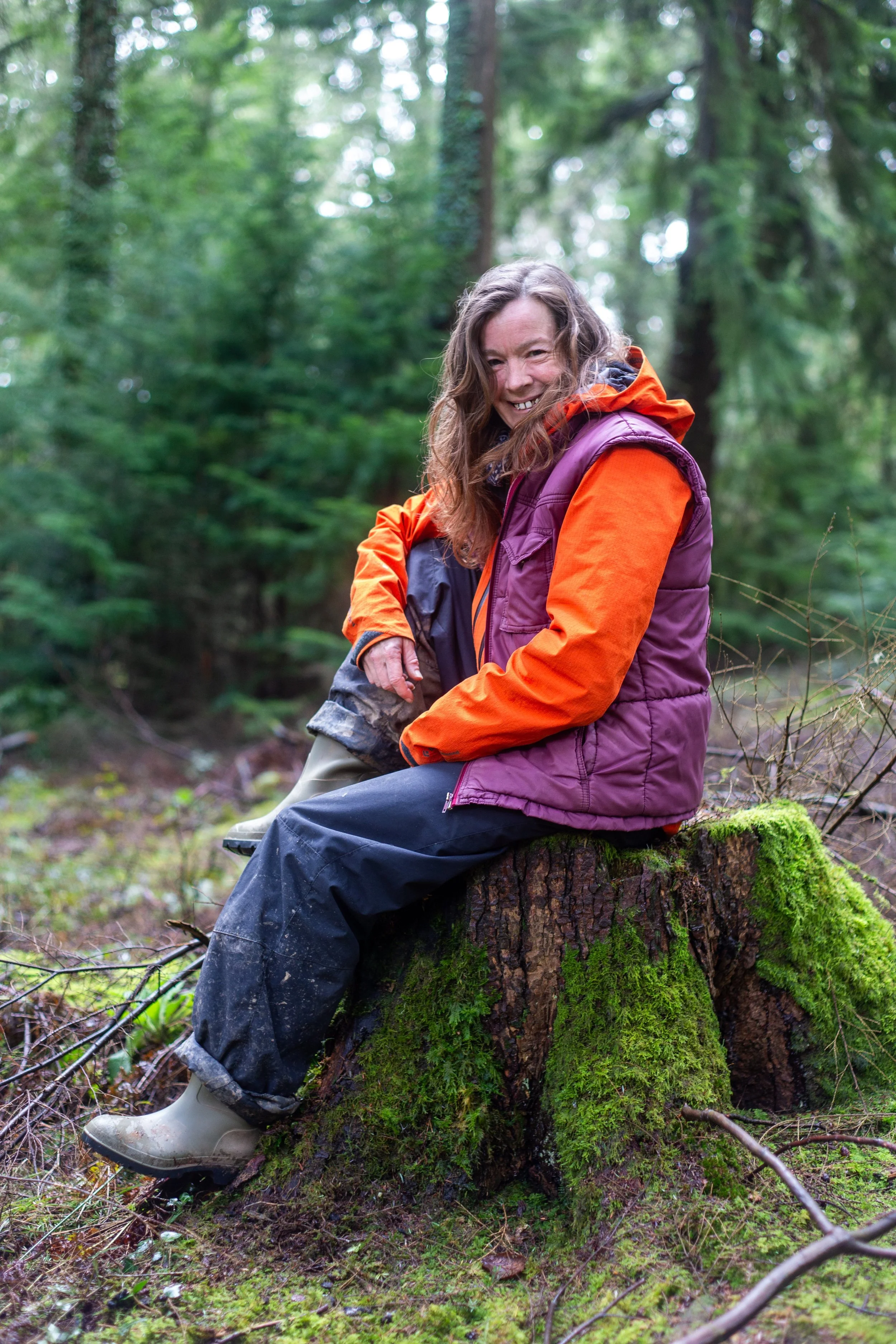 A woman sitting on a moss-covered tree stump in a forest, smiling and wearing outdoor clothing including a purple vest, orange jacket, and rubber boots.