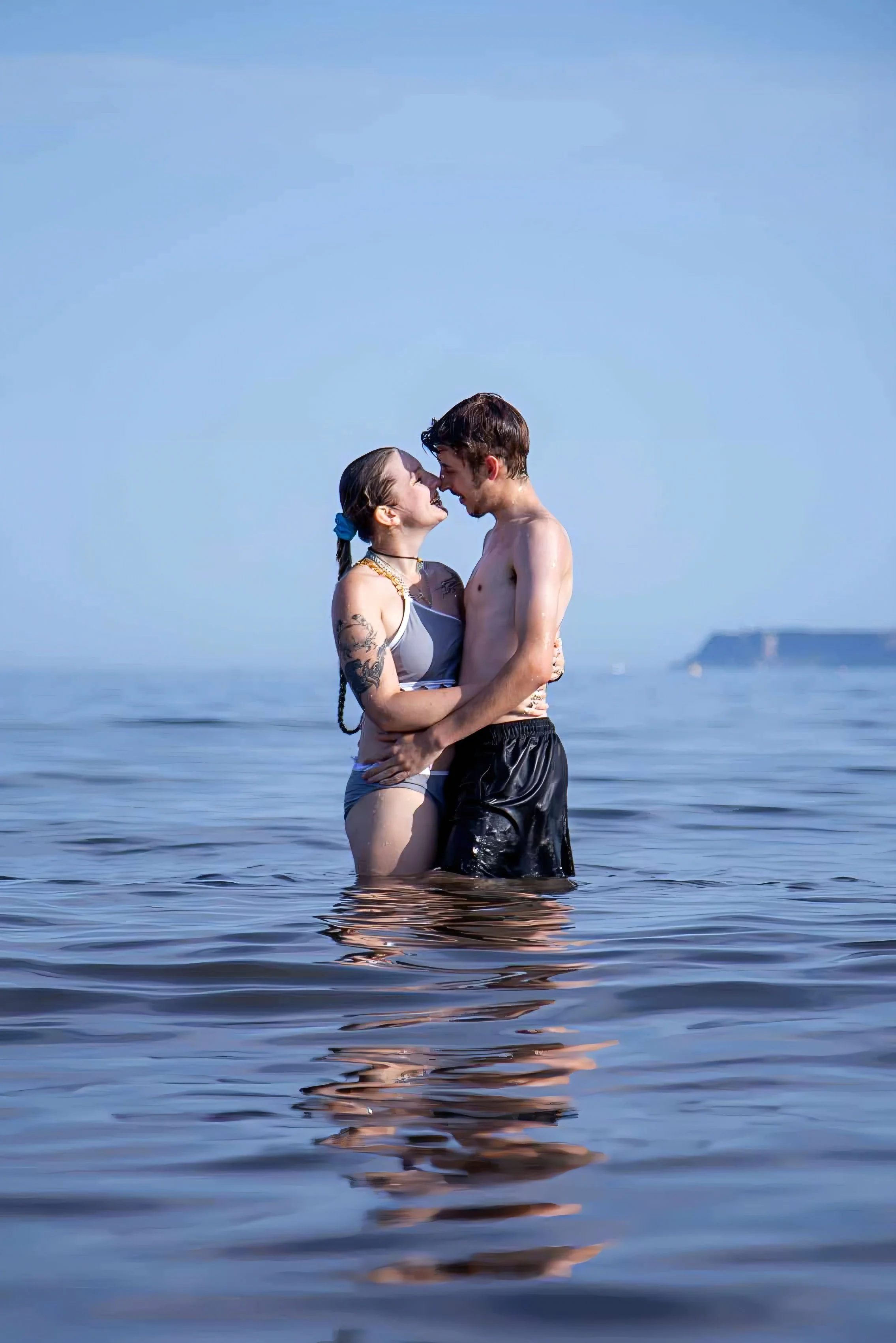 A couple stands in the ocean, embracing and smiling at each other during daylight with clear blue skies, with a distant coastal cliff on the horizon.