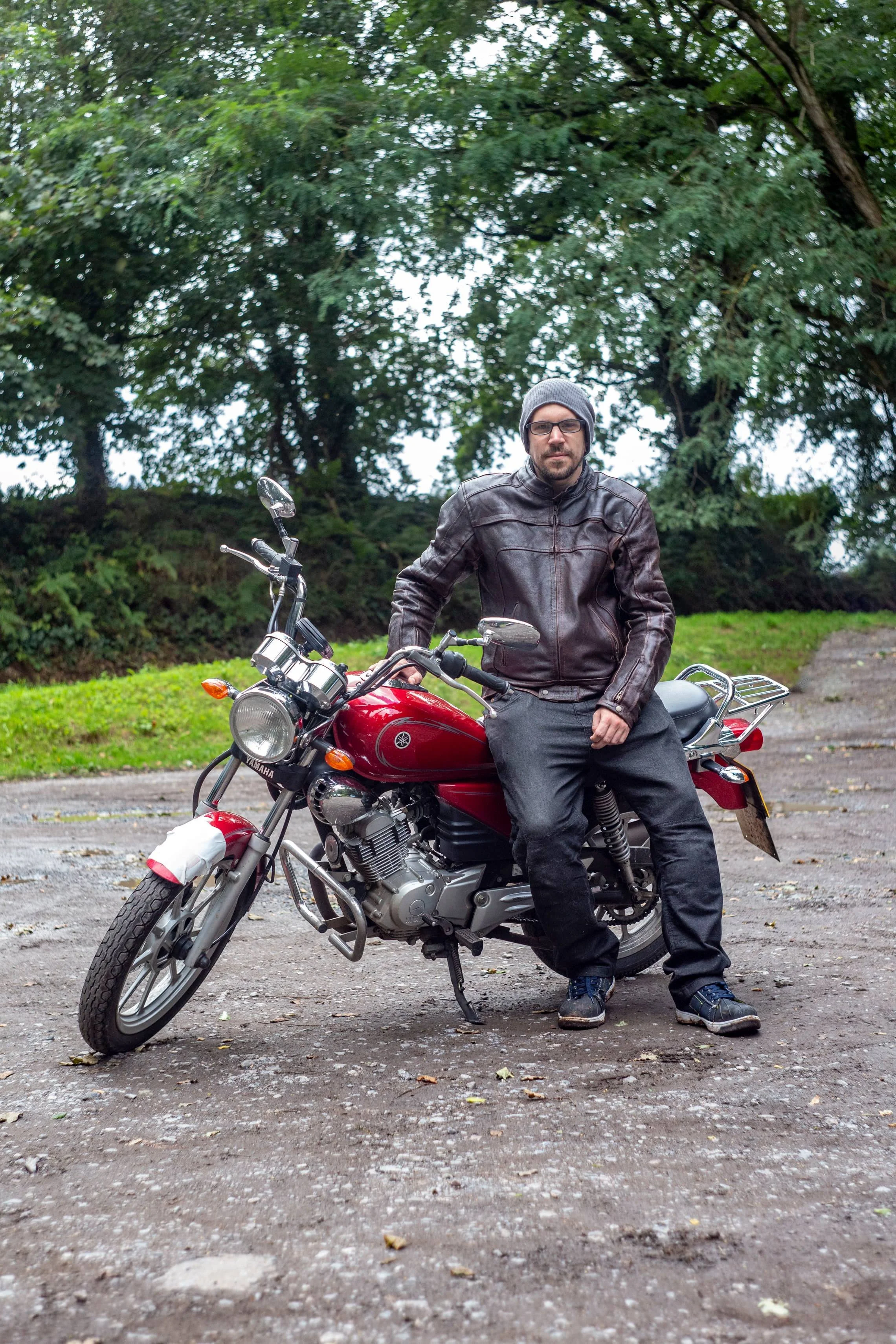 Man wearing a leather jacket, beanie, and jeans sitting on a red Yamaha motorcycle outdoors on a dirt surface, with green trees in the background.