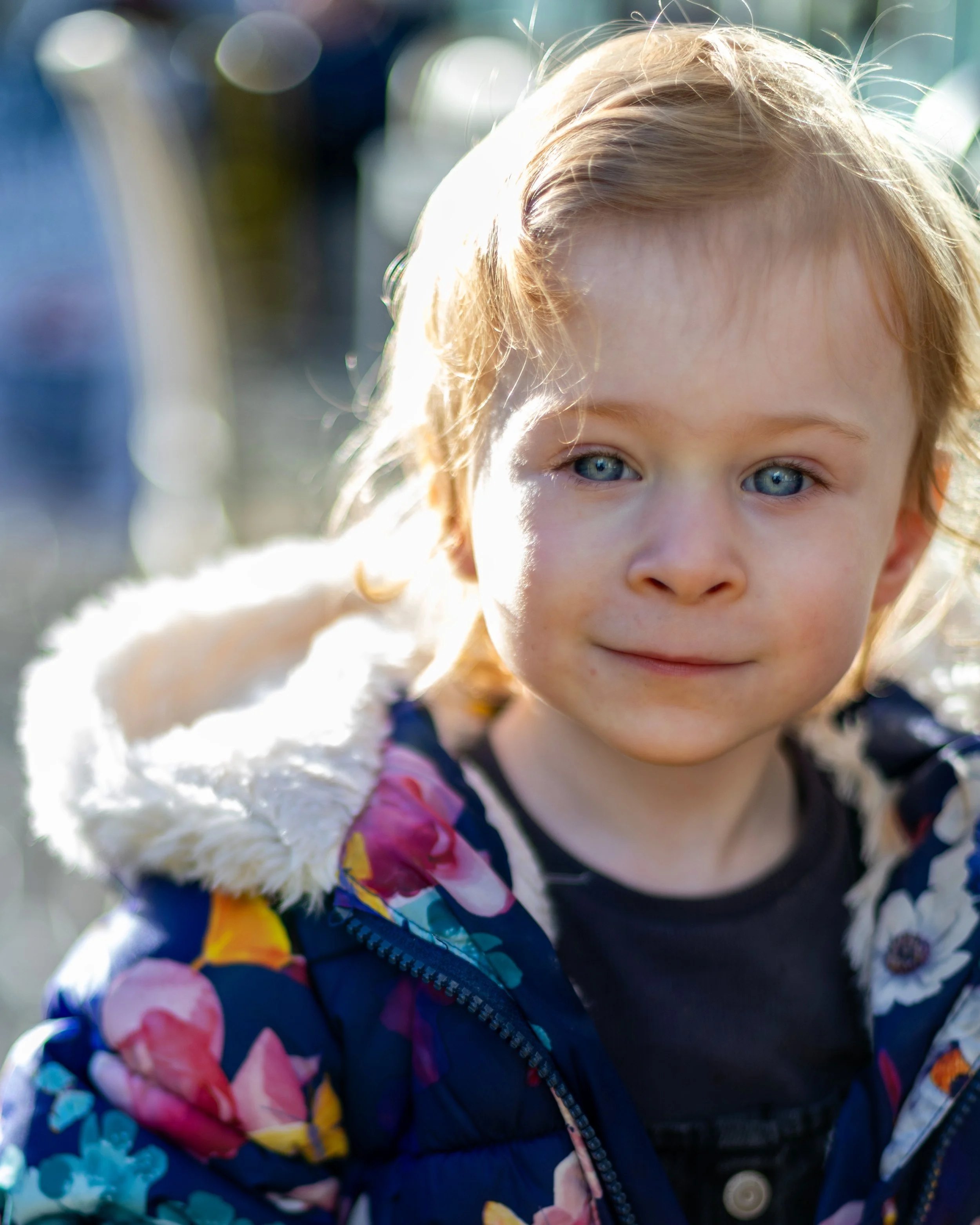 Close-up of a young child with blue eyes and blonde hair, wearing a dark floral jacket with a white furry hood, outdoors in sunlight.