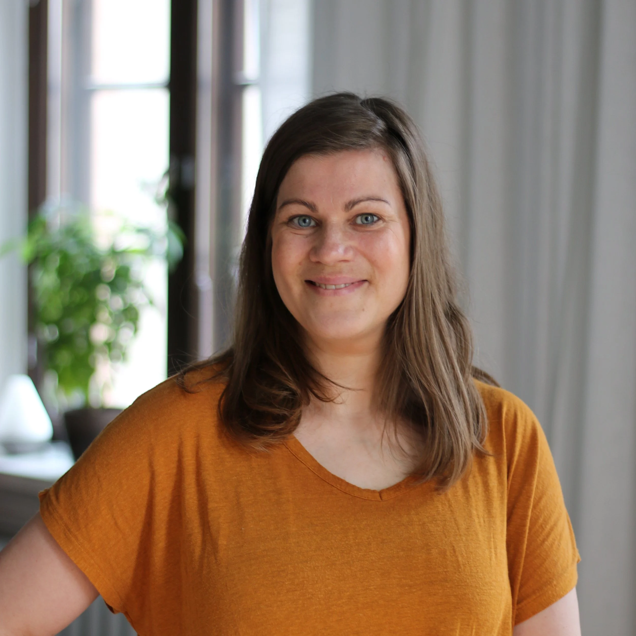 A woman with shoulder-length brown hair and blue eyes, wearing a mustard-colored top, smiling in a well-lit room with large windows and greenery in the background.