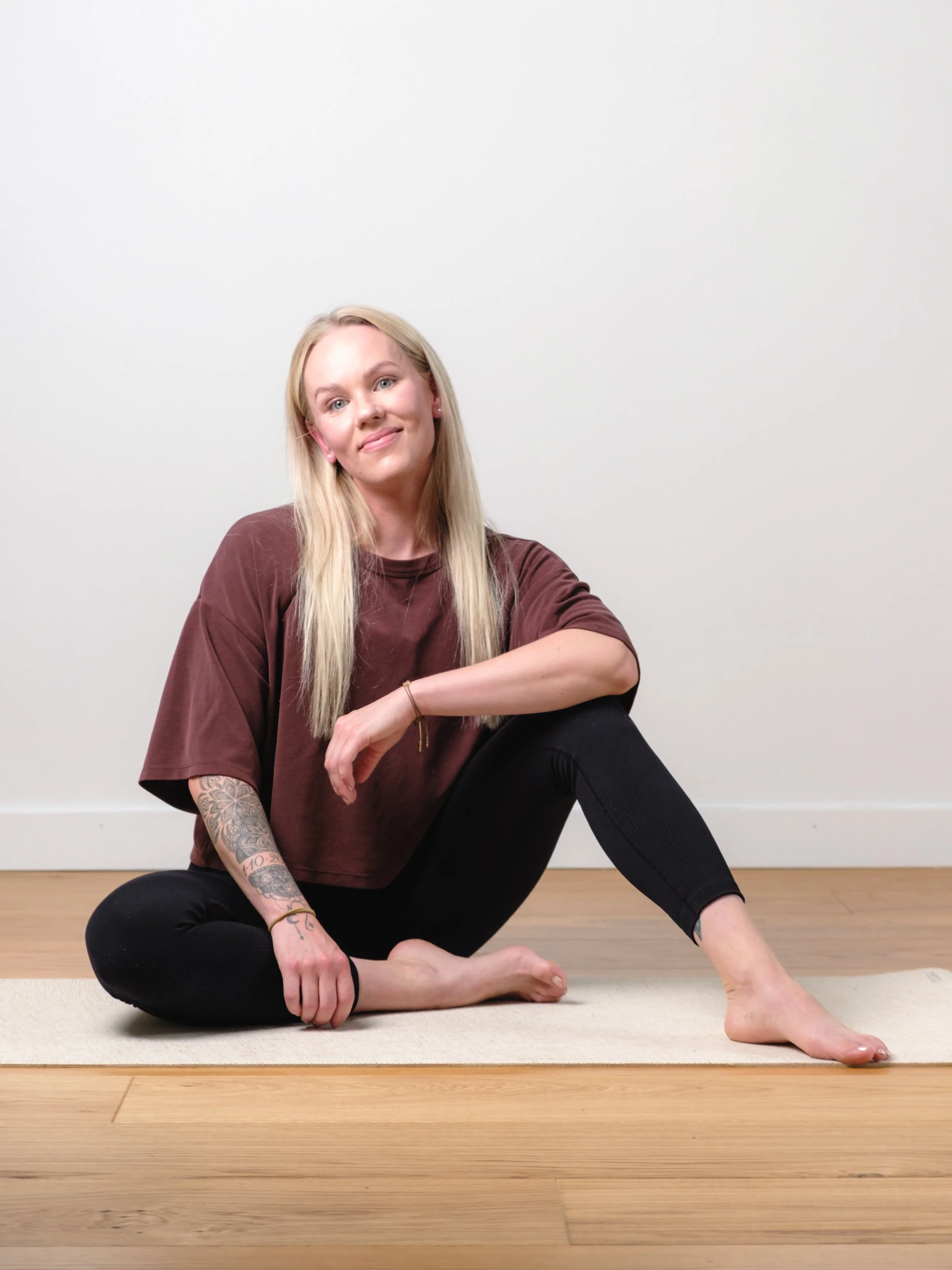 A blonde woman with blue eyes wearing a black athletic top with the logo 'ICIW', standing indoors with arms crossed, smiling softly, in front of a window with plants and white curtains.