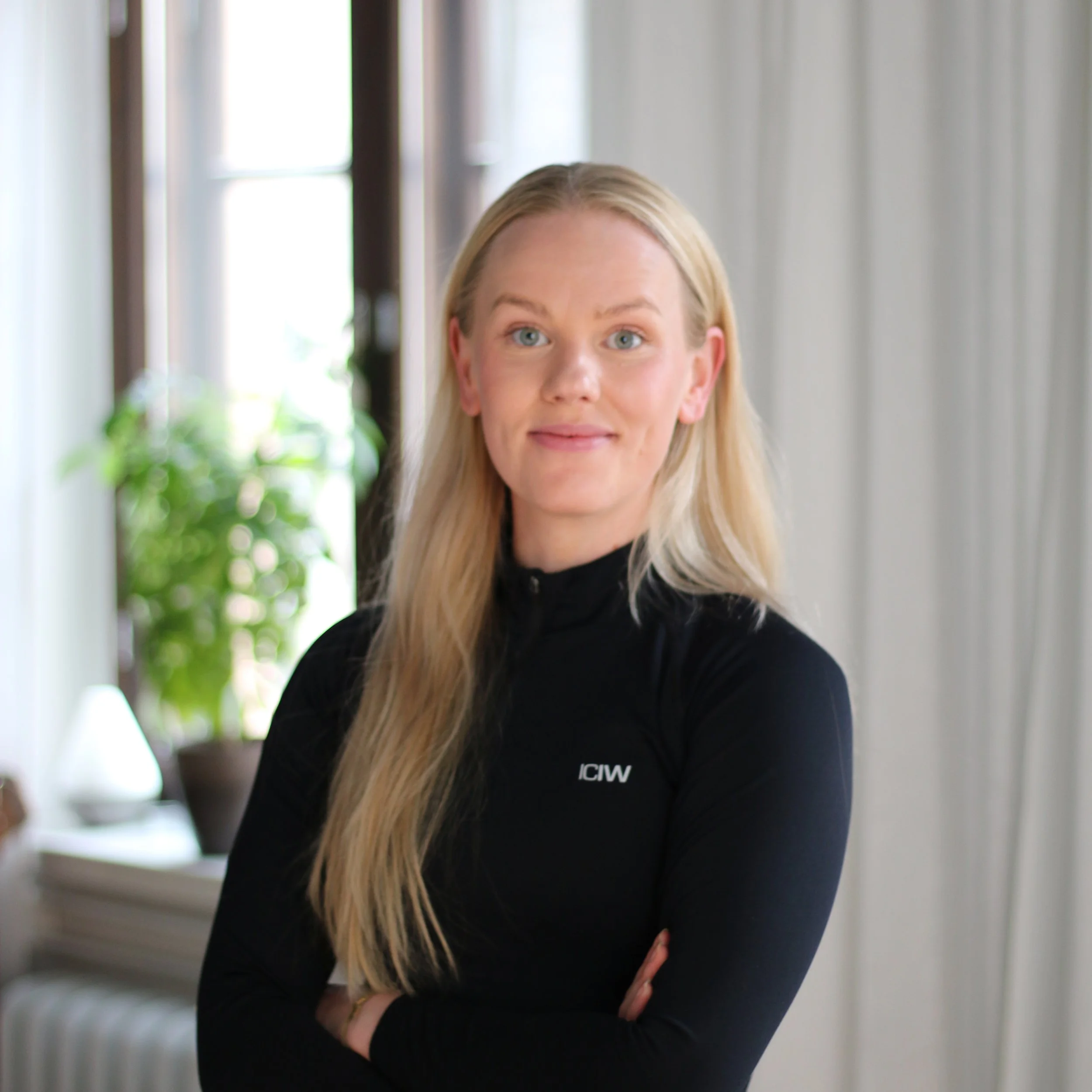 A blonde woman with blue eyes wearing a black athletic top with the logo 'ICIW', standing indoors with arms crossed, smiling softly, in front of a window with plants and white curtains.