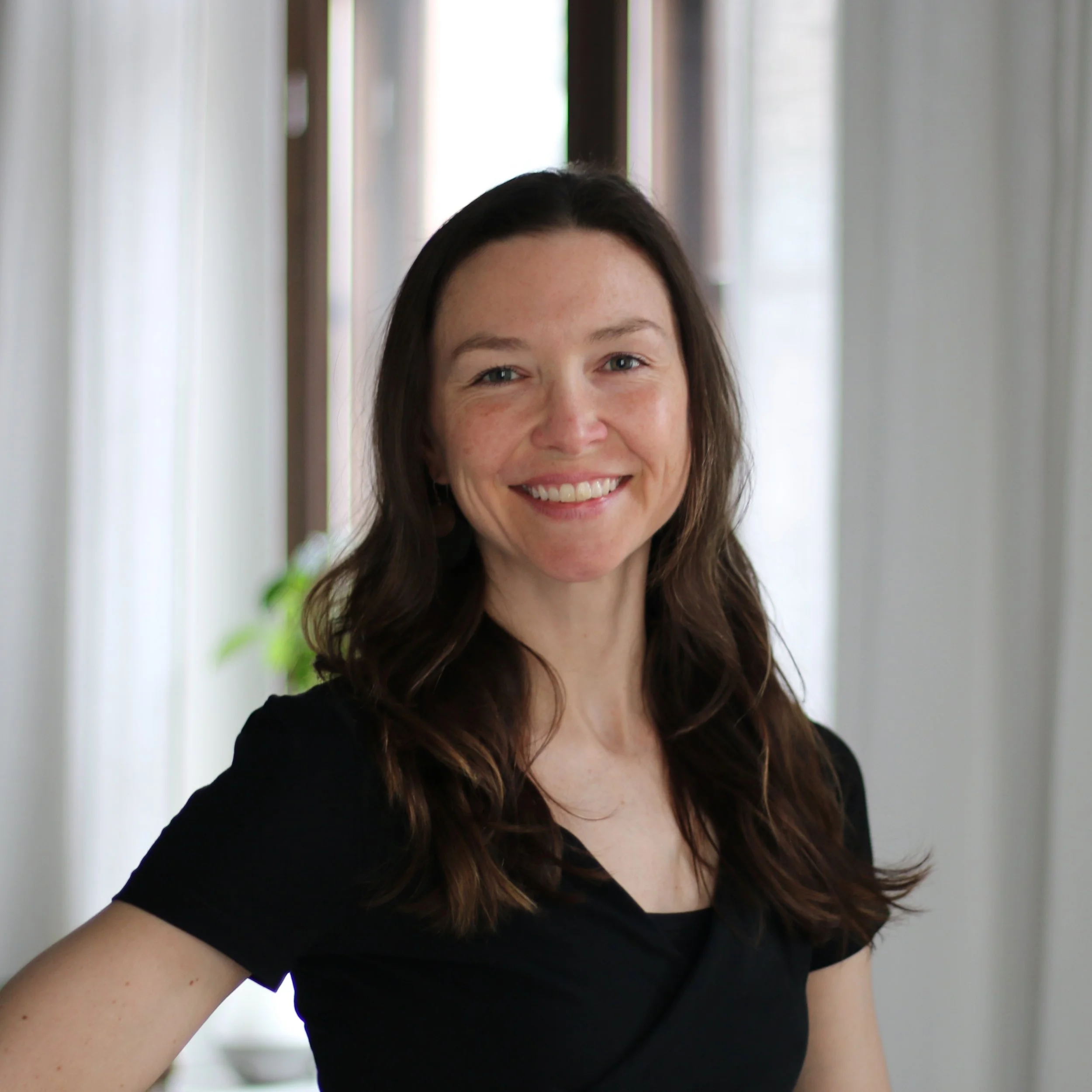 A woman with long dark hair smiling in a well-lit room with large windows and white curtains.