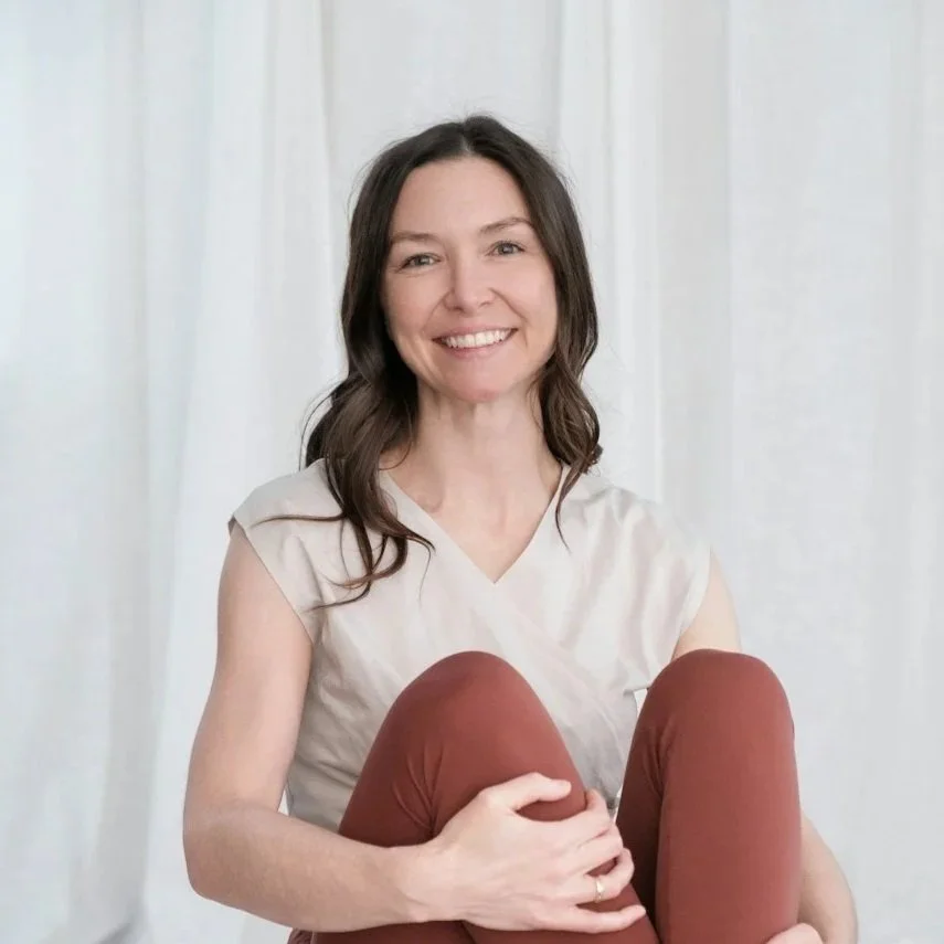 A woman with long dark hair smiling in a well-lit room with large windows and white curtains.
