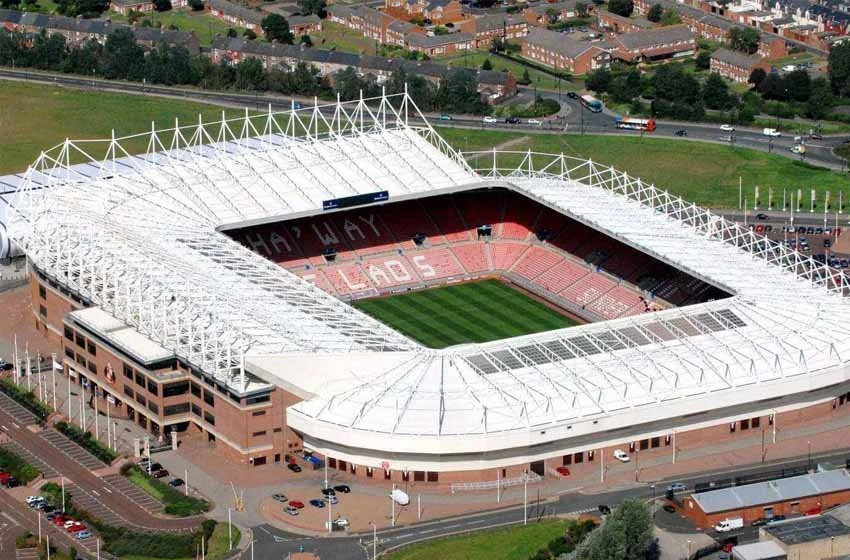 Aerial view of a football stadium with a white roof and green field, surrounded by a parking lot and cityscape in the background.