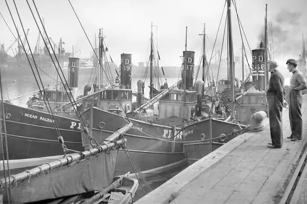 Black and white photo of several vintage boats docked at a pier, with a couple of men standing on the dock observing.