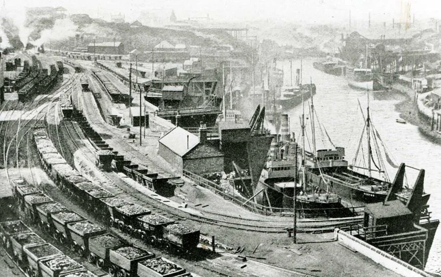 Historical black-and-white photo of a busy port with ships docked and numerous train tracks in the foreground.