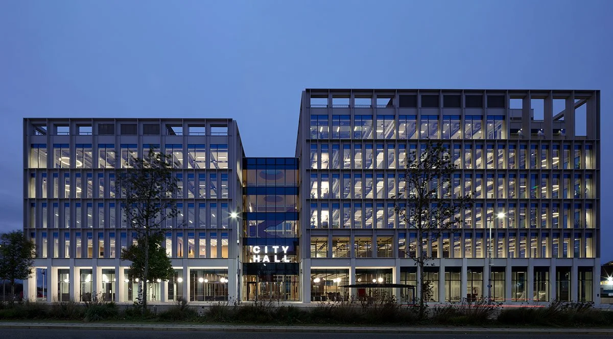 Modern multi-story city hall building at dusk with illuminated windows and a central glass entrance.