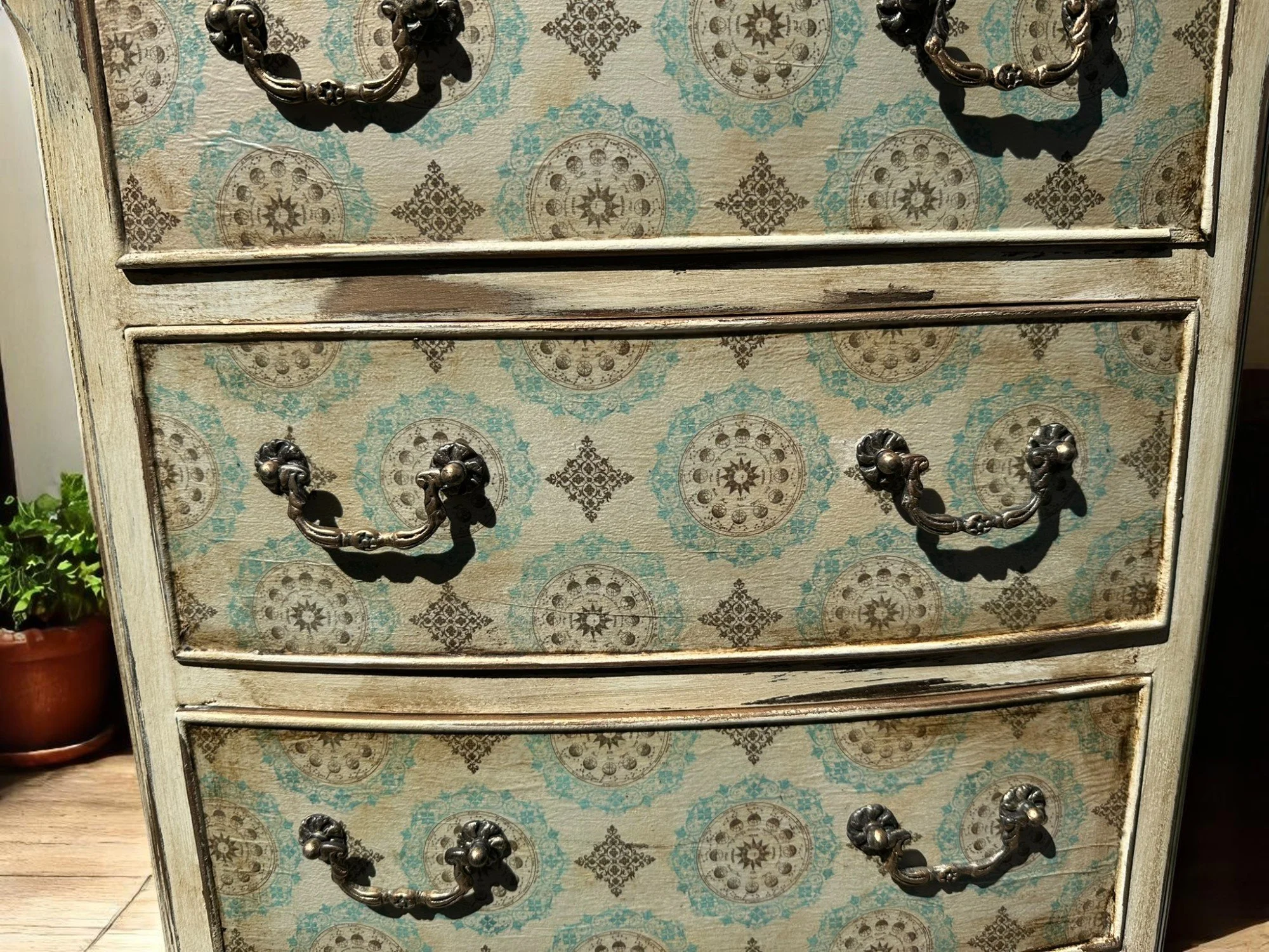 A vintage wooden dresser with three drawers, decorated with a patterned paper surface featuring blue and gray circular and diamond designs. The dresser has ornate metal handles on each drawer.