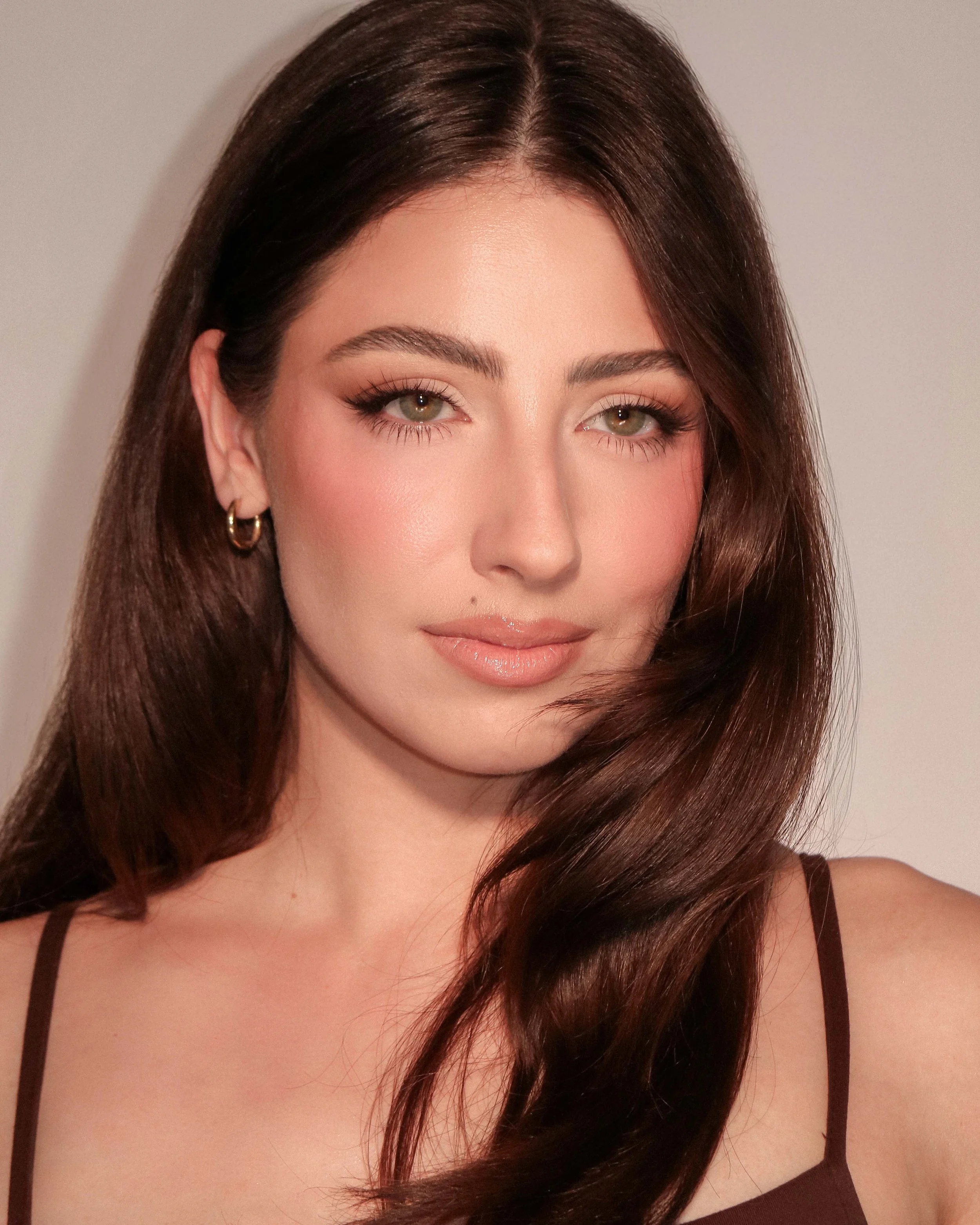 A young woman with long, wavy brown hair and light makeup, wearing a dark spaghetti strap top and gold hoop earrings, looking into the camera against a plain background.