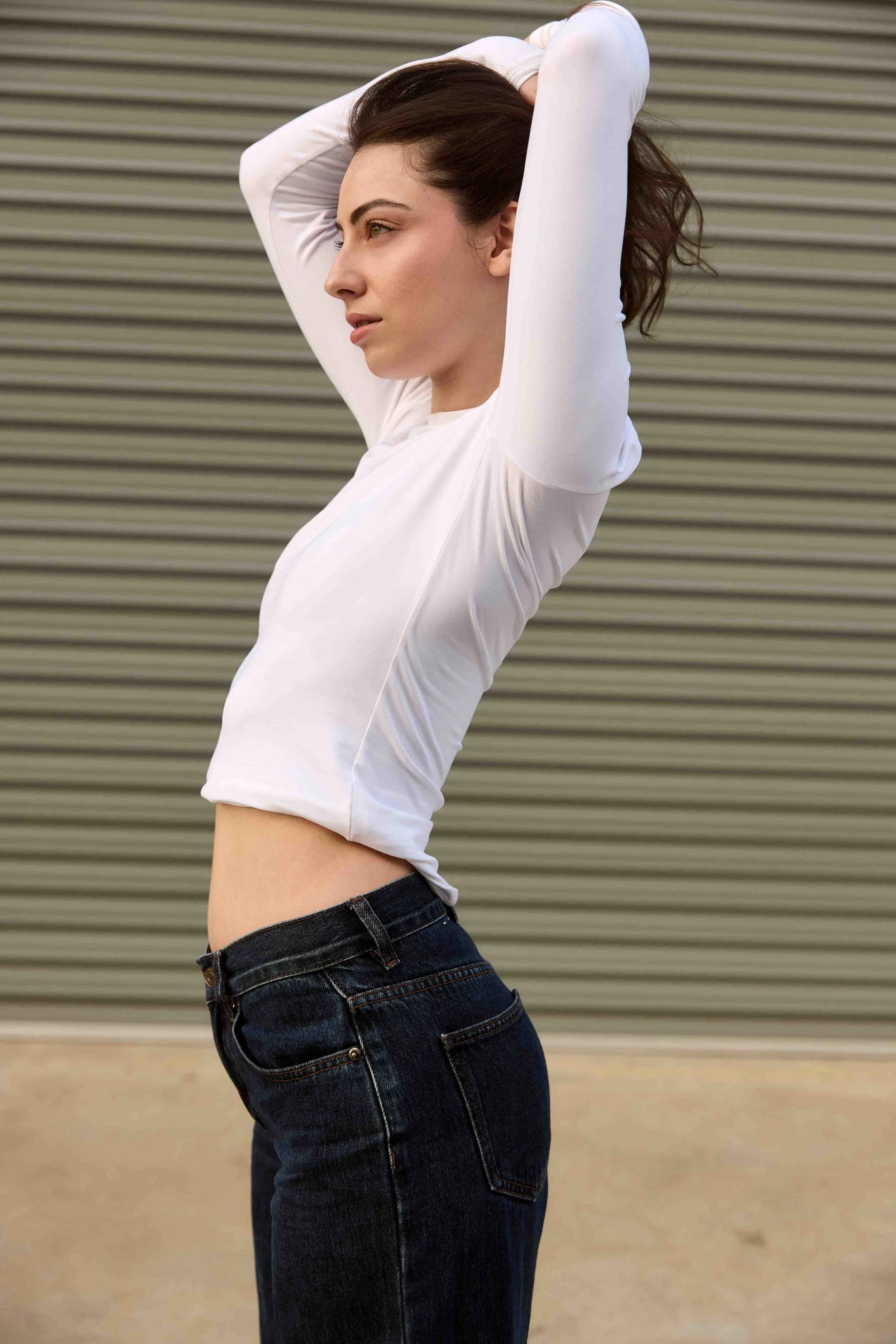 A woman with brown wavy hair wearing a white long-sleeve top and dark blue jeans standing against a gray industrial background with horizontal ridges, stretching her arms above her head.