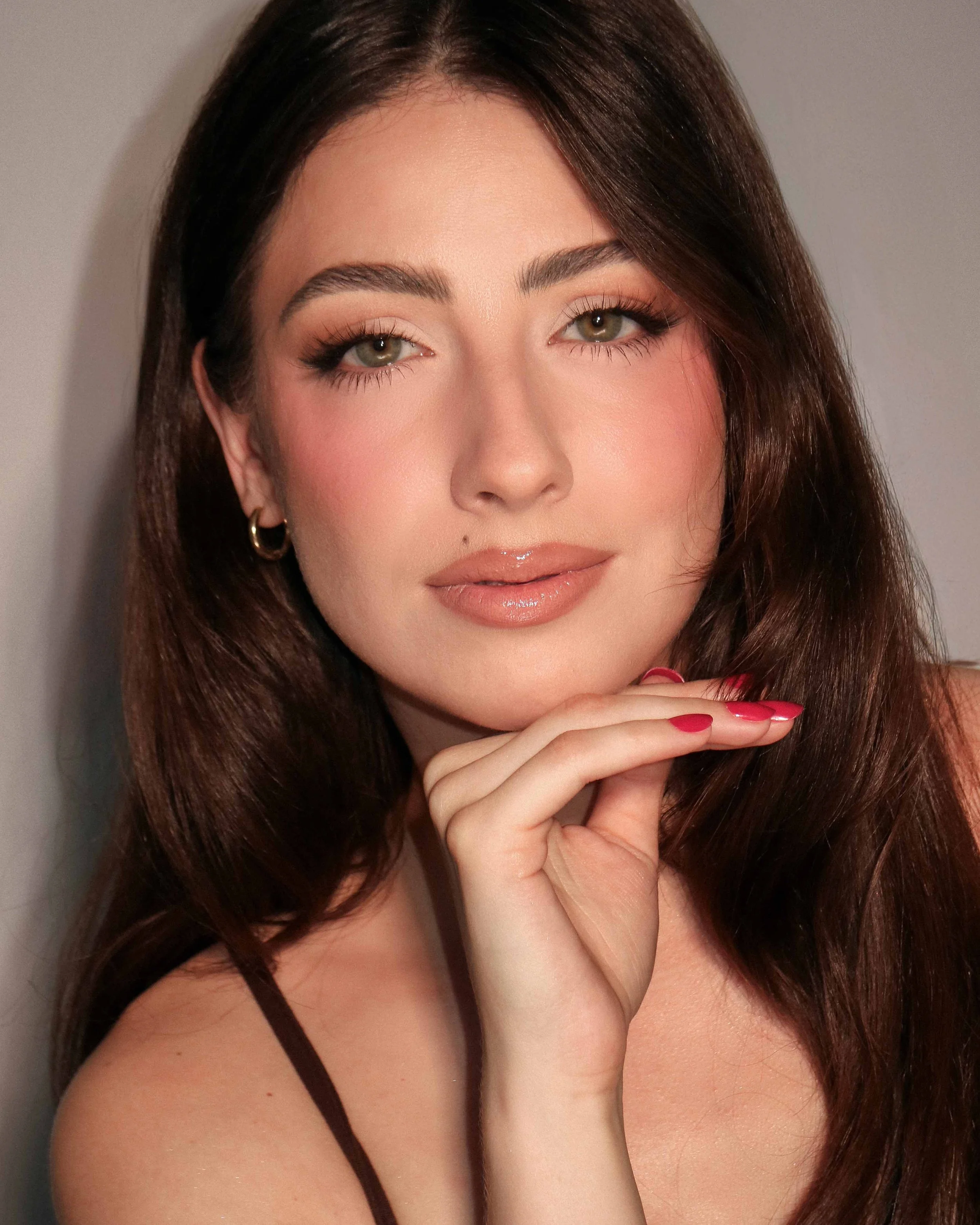 Close-up portrait of a woman with long dark brown hair, light makeup, wearing earrings, and a spaghetti strap top, with her hand resting near her chin.