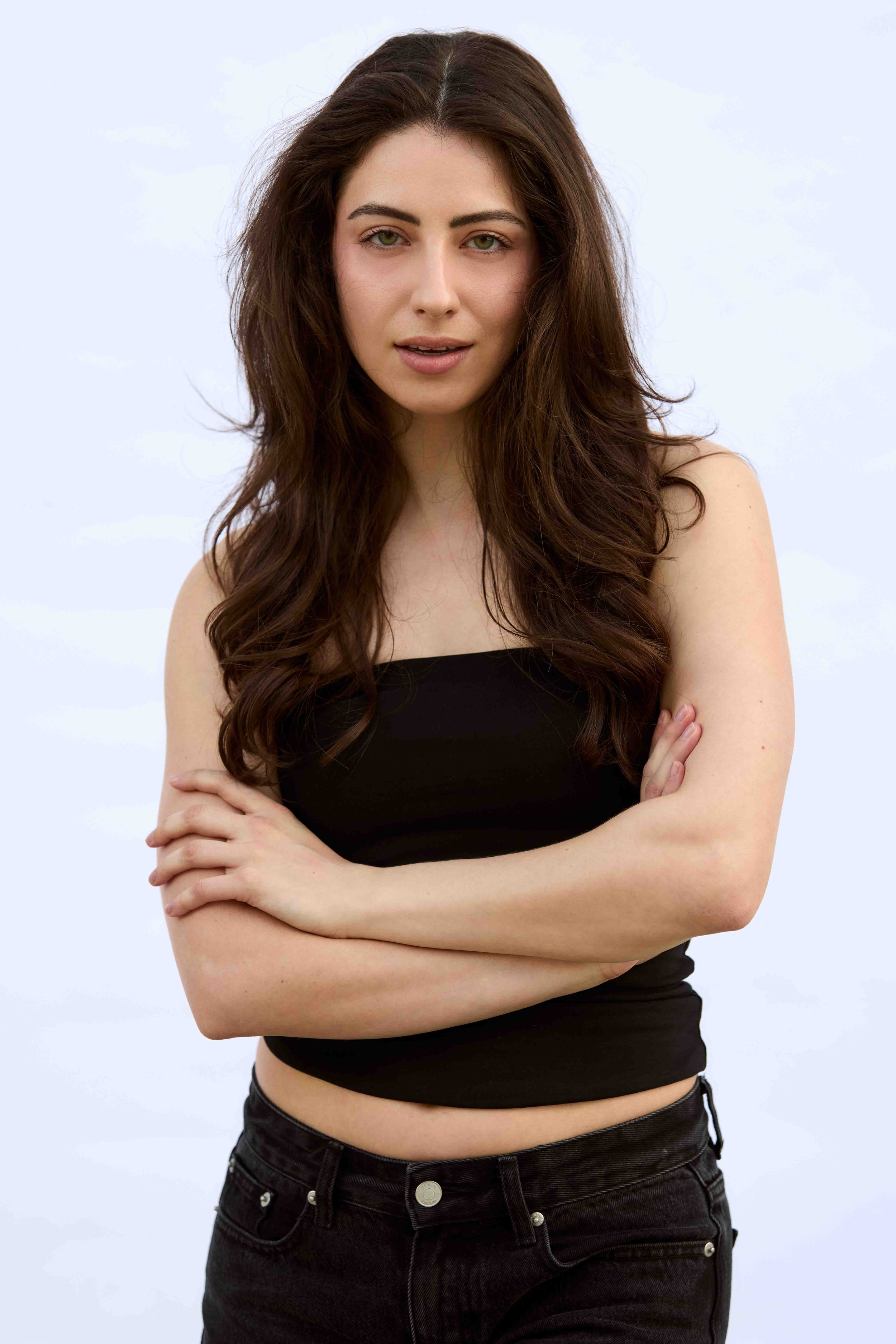 A woman with long, wavy brown hair and light skin, standing with arms crossed, wearing a black sleeveless top and black jeans, against a light blue background.
