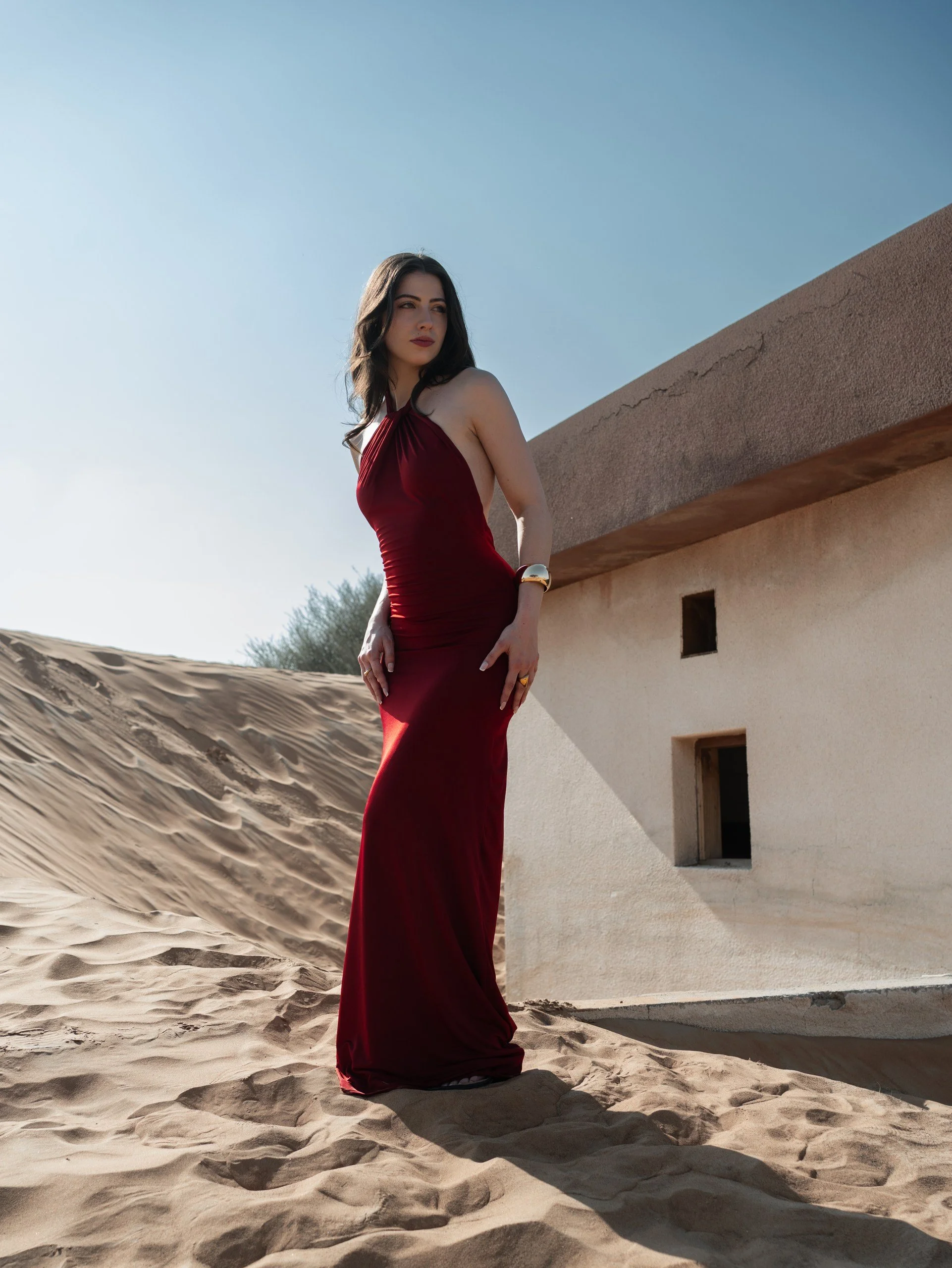 A woman in a long, red dress standing on sand dunes next to a beige stone building with small windows.