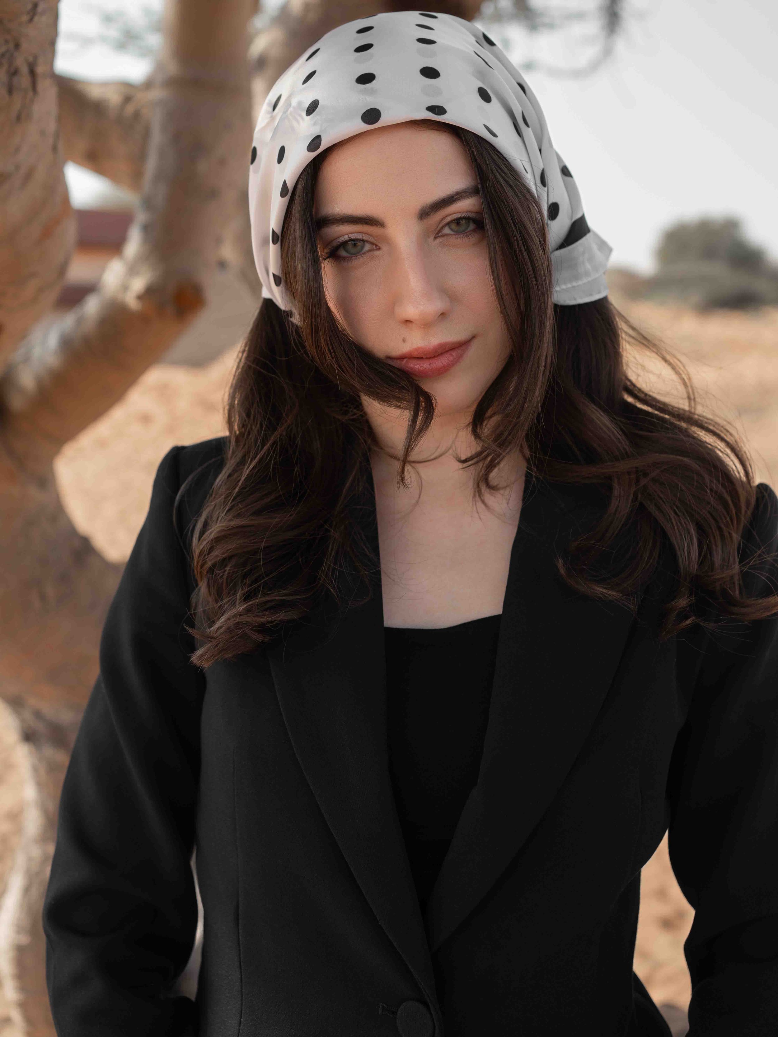 A woman with long dark hair wearing a white headscarf with black polka dots, a black blazer, and a black top, standing outdoors with a desert landscape and a tree in the background.
