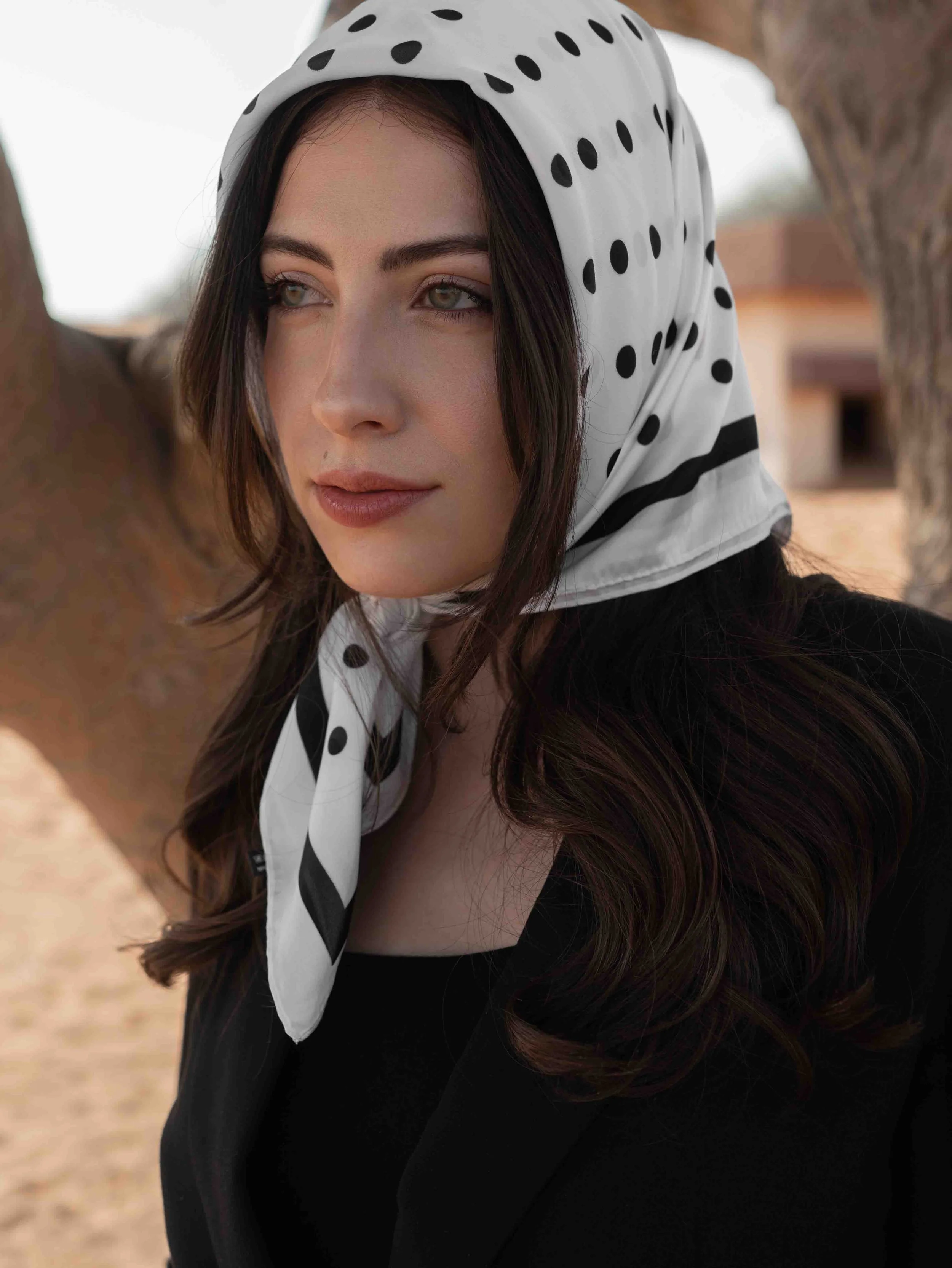 A woman with dark hair and fair skin wearing a black top and blazer, with a white polka dot scarf tied around her head, standing outdoors near a tree with a building in the background.