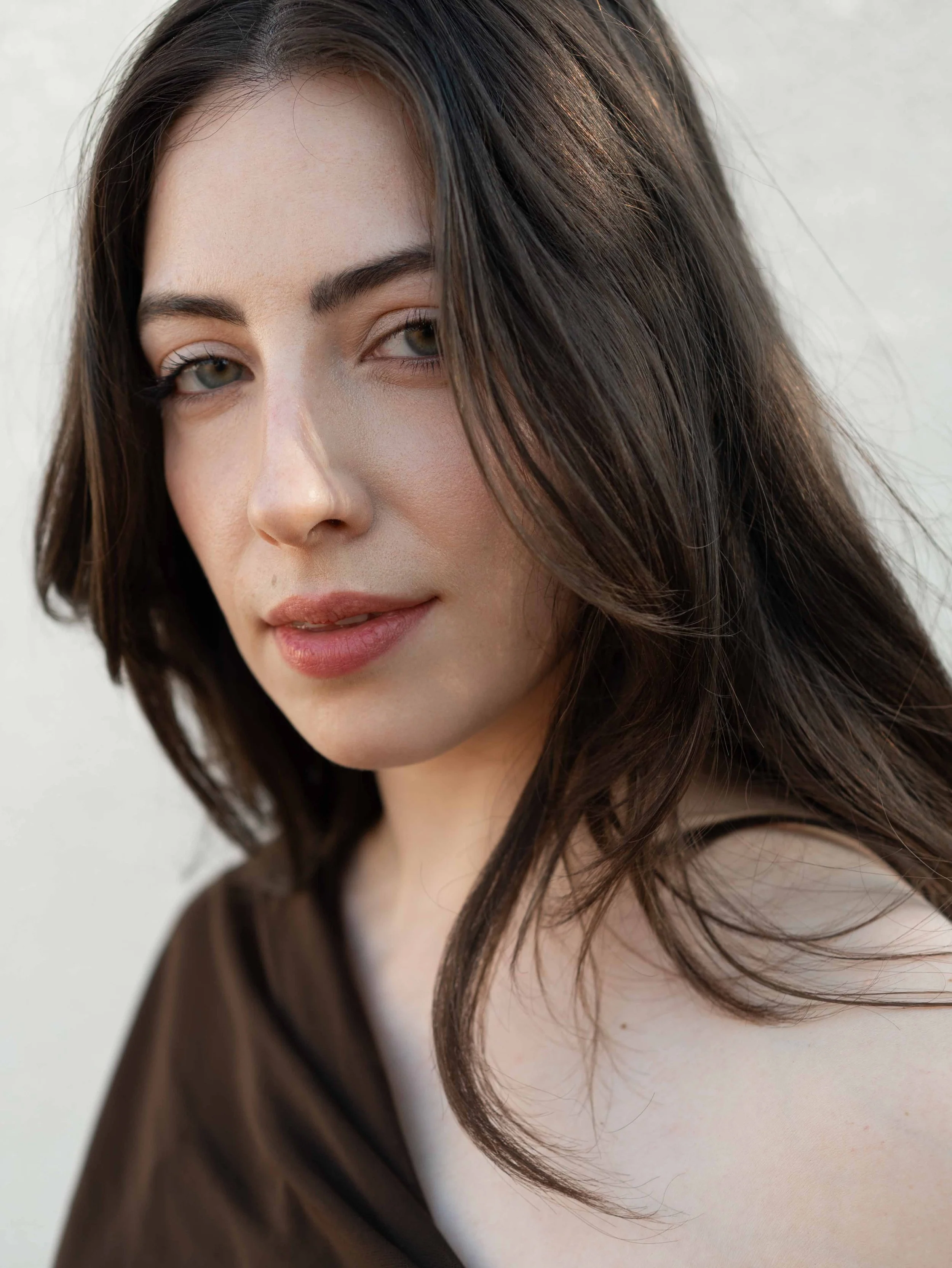 Close-up of a woman with long wavy brown hair, light makeup, and a subtle smile, looking at the camera against a plain background.