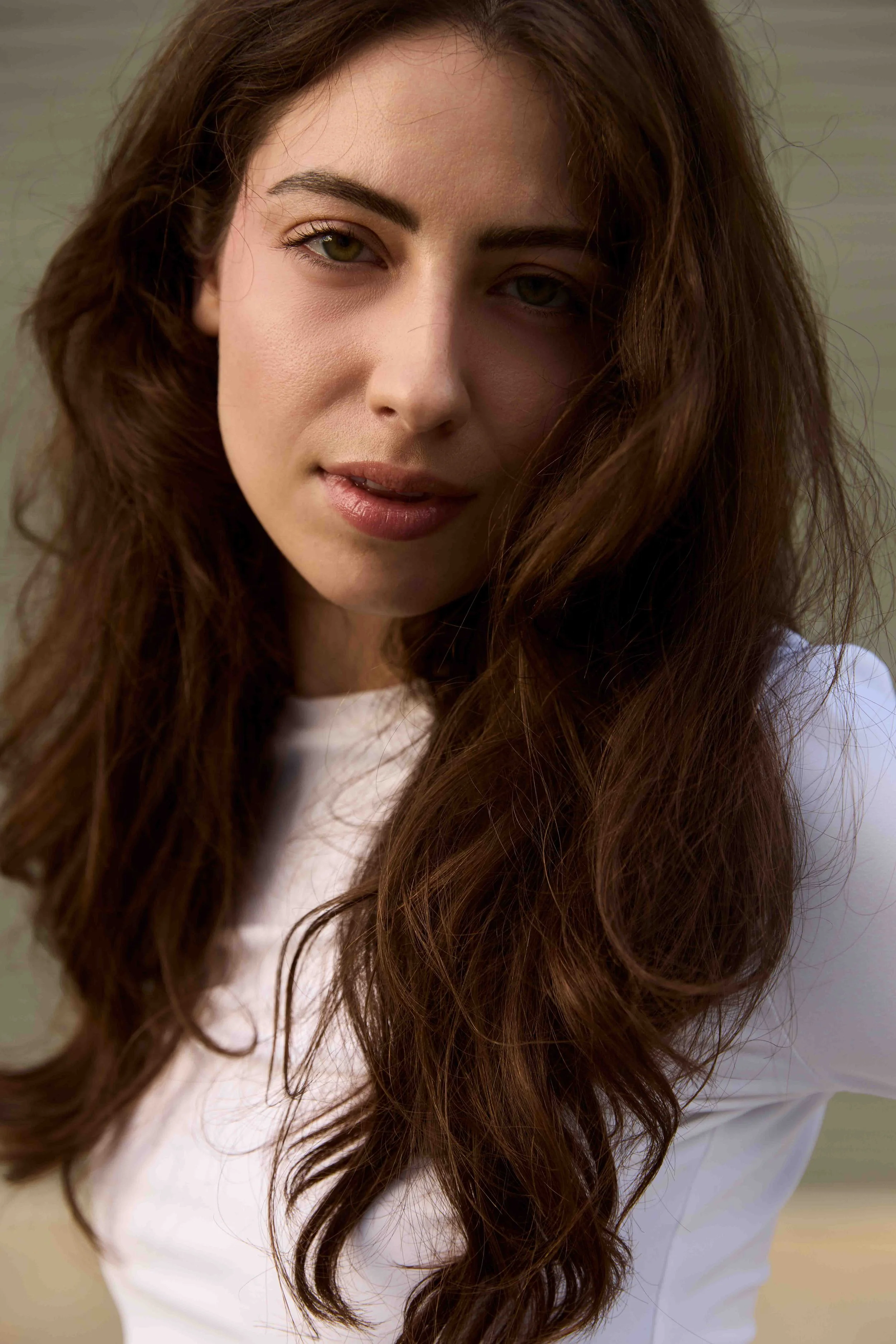 Close-up of a young woman with long, wavy brown hair, wearing a white top, looking at the camera with a slight smile.