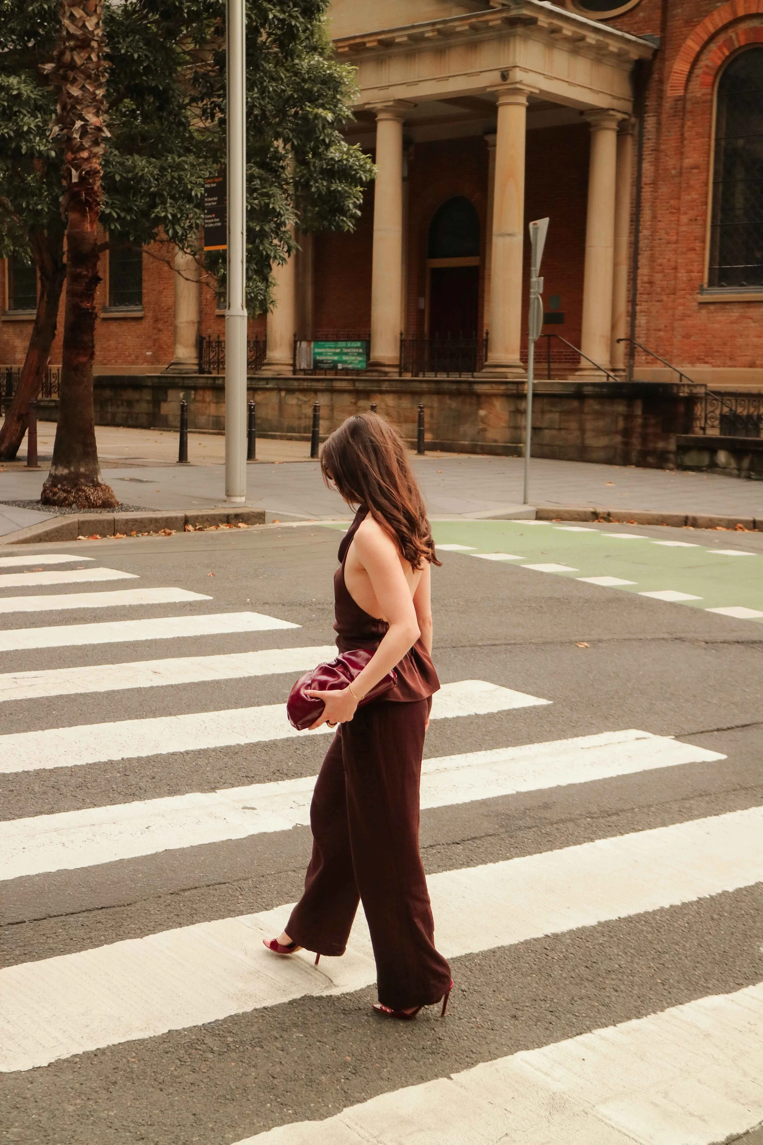 A woman dressed in dark clothing and high heels crossing a pedestrian crosswalk on a city street, holding a maroon purse.