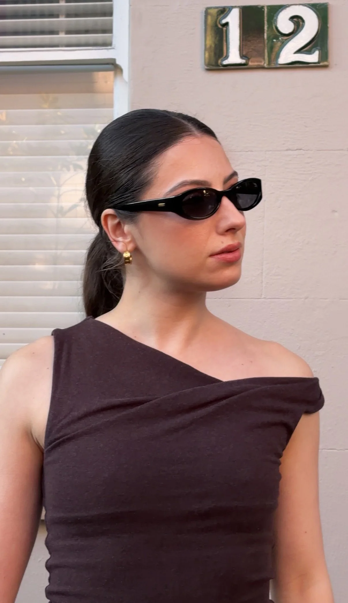 A woman with dark hair pulled back, wearing black sunglasses, gold hoop earrings, and a dark off-shoulder top, standing outside near a beige wall with house numbers '112' on the wall.