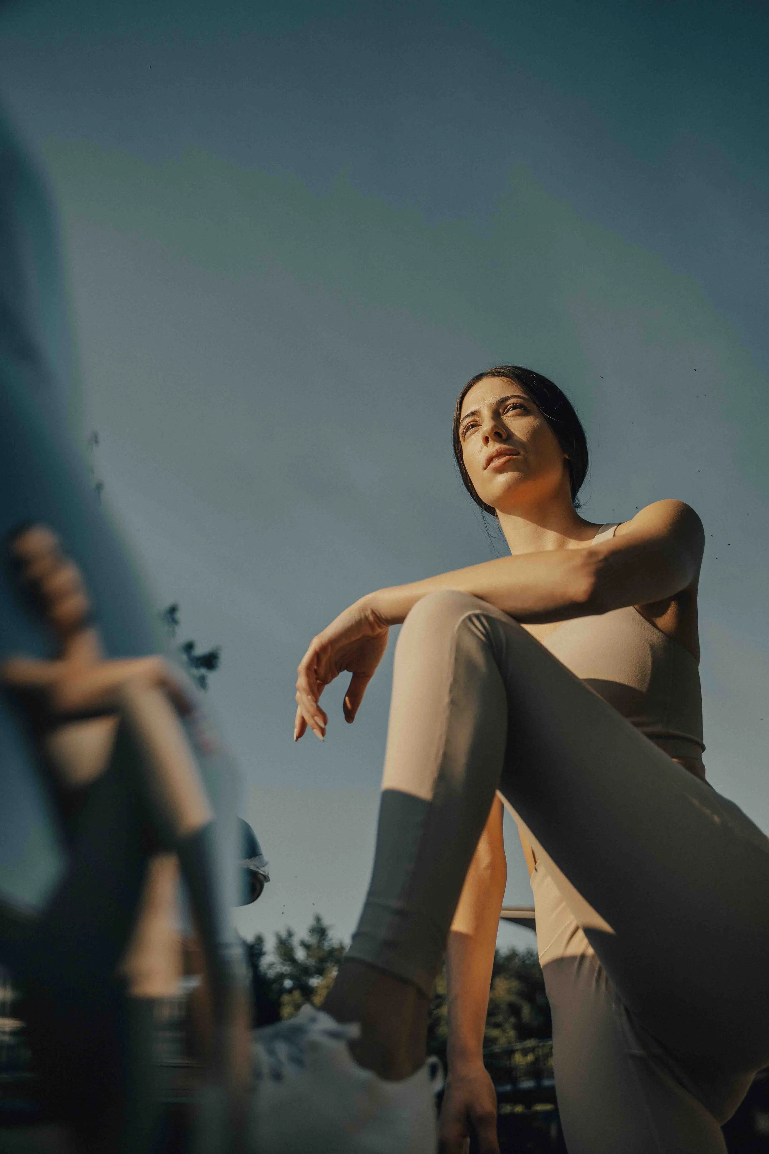 Low-angle photograph of a woman in athletic wear sitting outdoors, looking into the distance with a thoughtful expression under a clear sky, with blurred people and trees in the background.