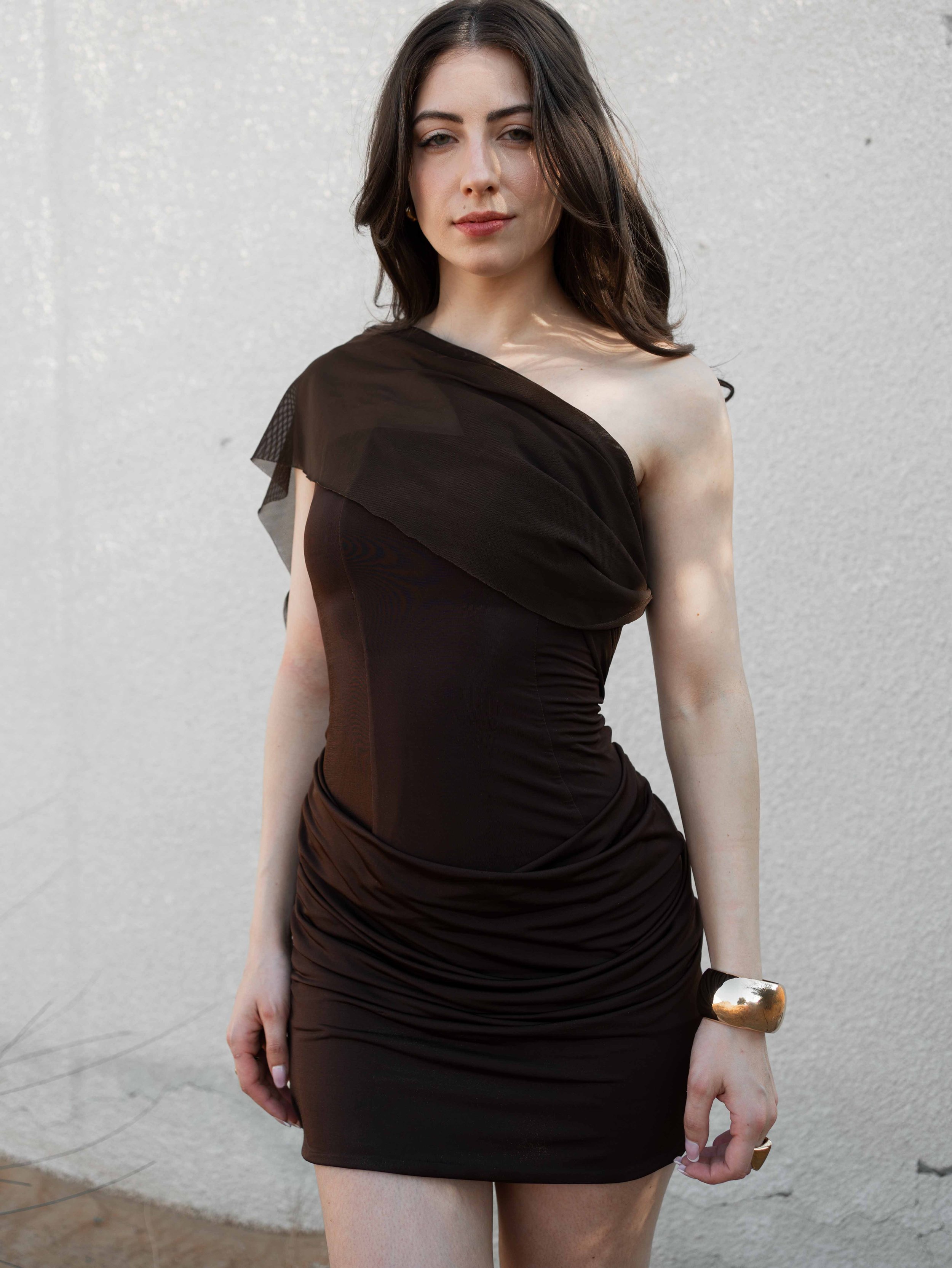 A woman with shoulder-length dark brown hair wearing a black dress with an asymmetrical sheer fabric detail on one shoulder and a gold bracelet on her wrist, standing against a plain white wall.