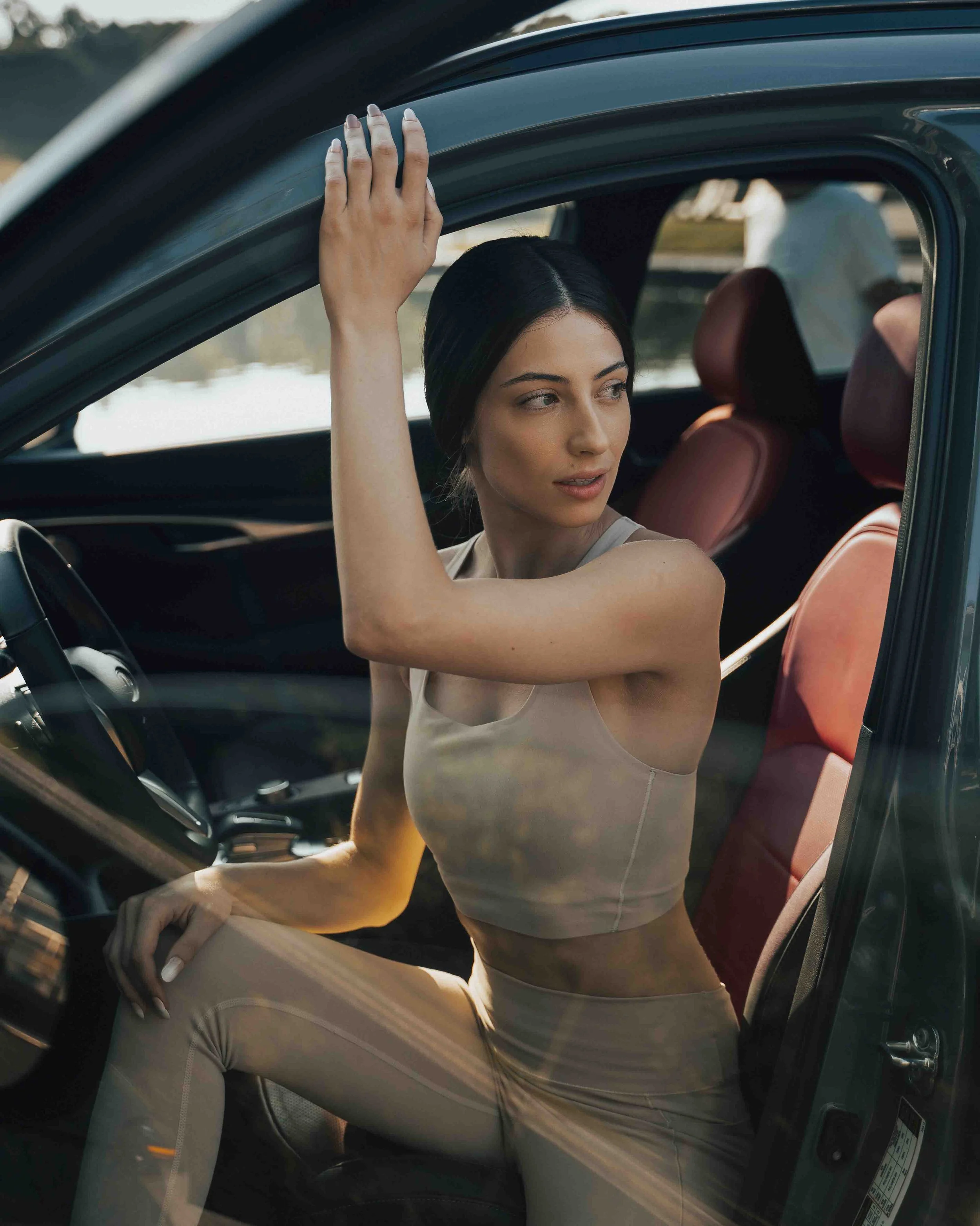 A woman with black hair and light skin sitting inside a car, looking out the window, with her arm resting on the window frame.