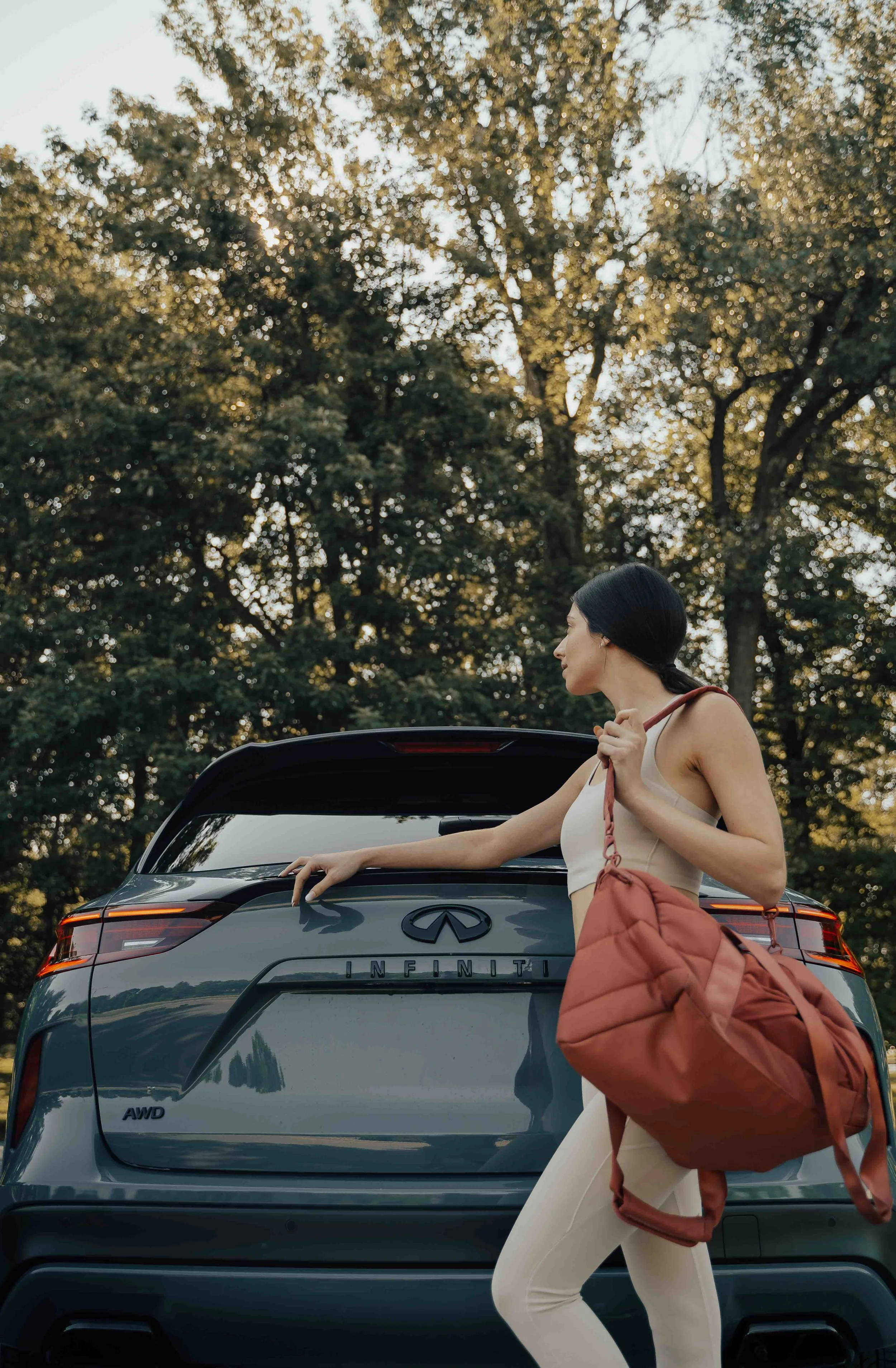 A woman with black hair and light skin standing beside the trunk of a gray Infiniti SUV with the logo and model tag visible. She is wearing a beige sports bra and cream-colored pants, carrying a large brown/pinkish bag slung over her shoulder. The ba